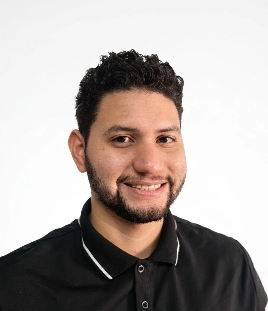 Portrait of a young man with dark, curly hair and a beard, smiling, wearing a black polo shirt with white trim against a plain white background.