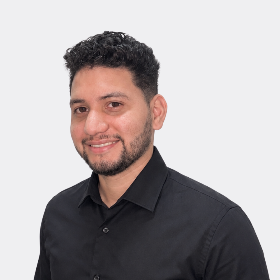 Portrait of a young man with dark, curly hair and a beard, smiling, wearing a black polo shirt with white trim against a plain white background.