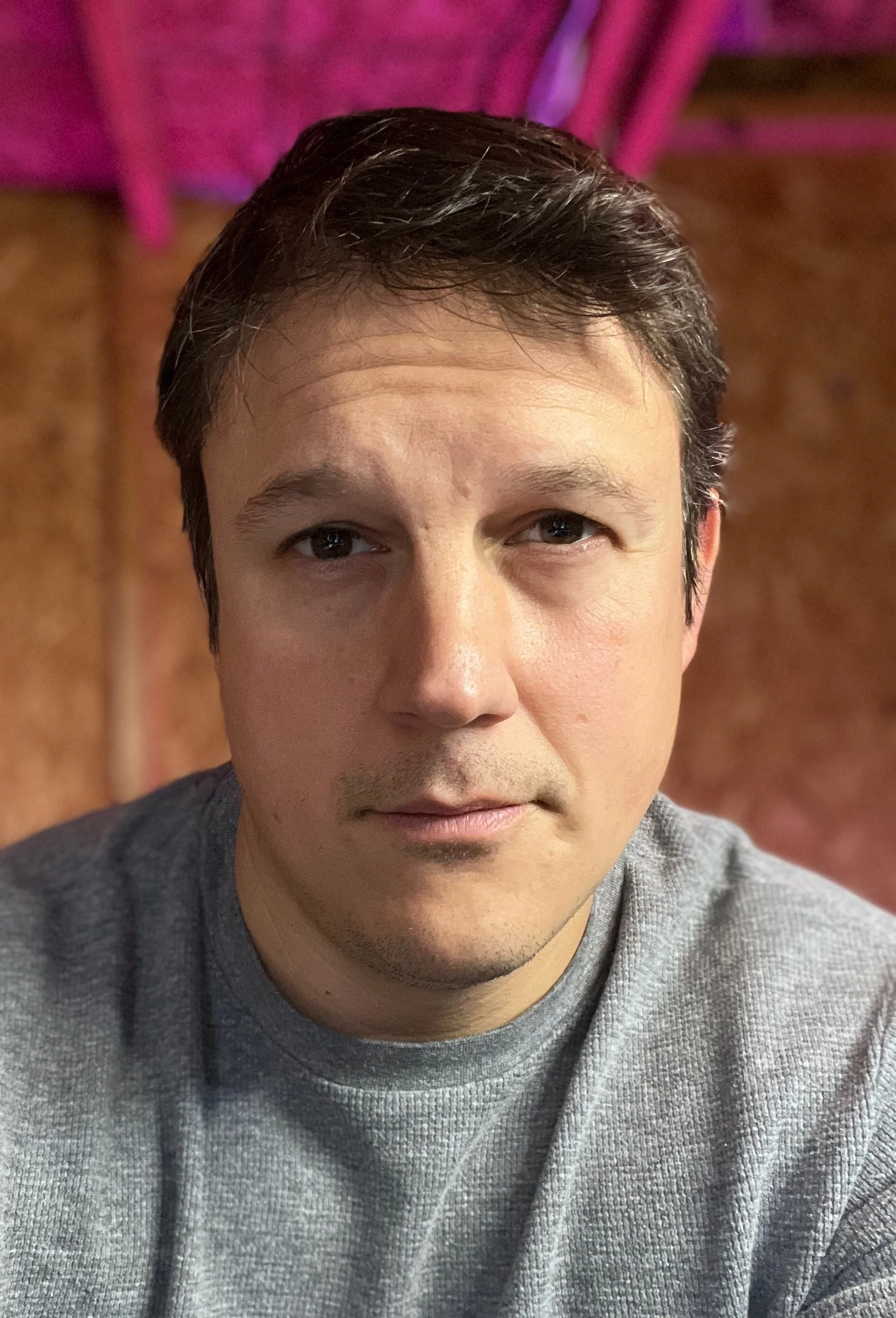 A close-up photo of a young man with short dark hair, beige skin, wearing a gray sweatshirt, looking into the camera with a neutral expression, inside a warmly lit indoor space.