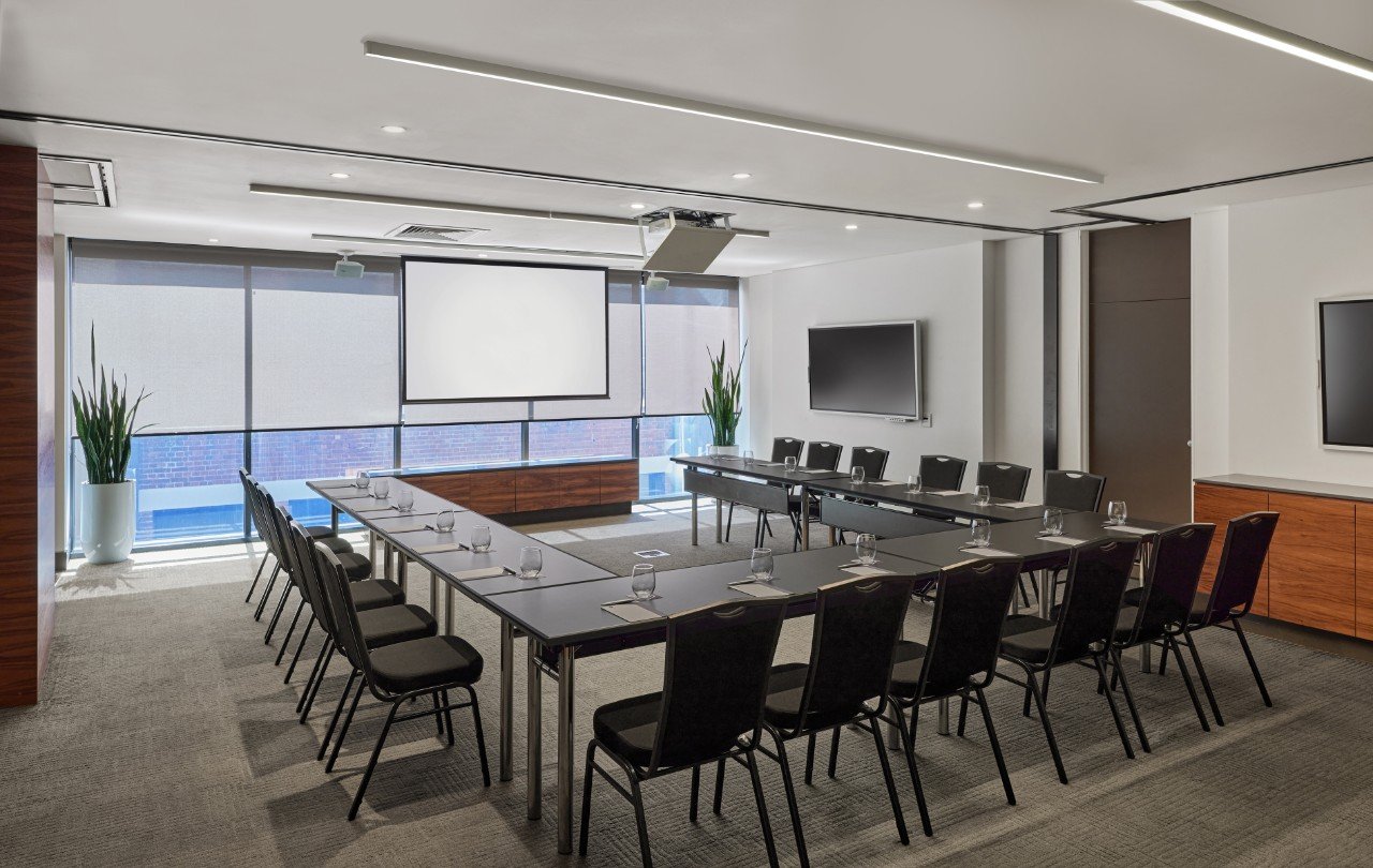 A set of desks are set up in a U shape with cups and paper on top, there is a projector set in front of it.