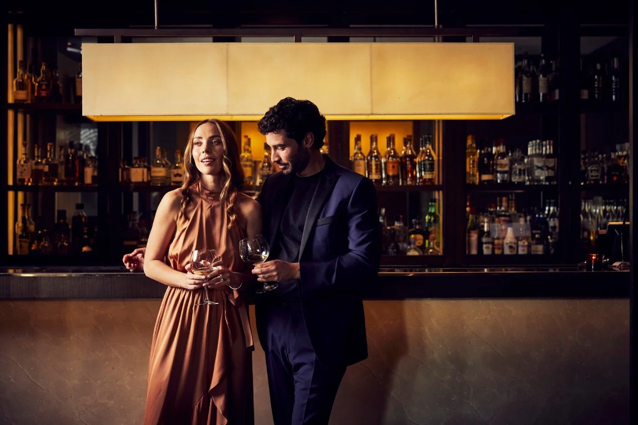 Woman and man stand at a bar with moody dark lighting, wearing formal attire and holding a glass of wine each.