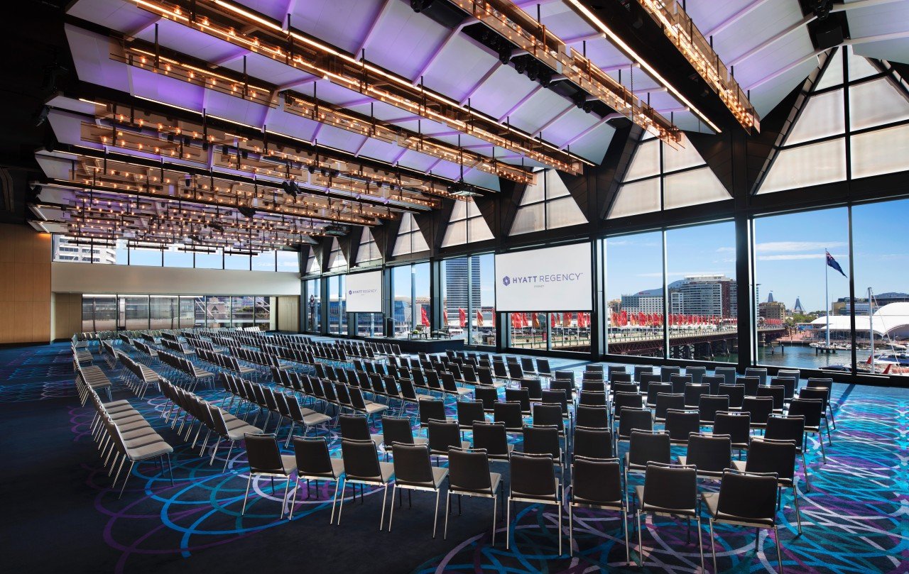 Large auditorium with seats set up theatre style, the view of darling harbour outside the windows on the far side.