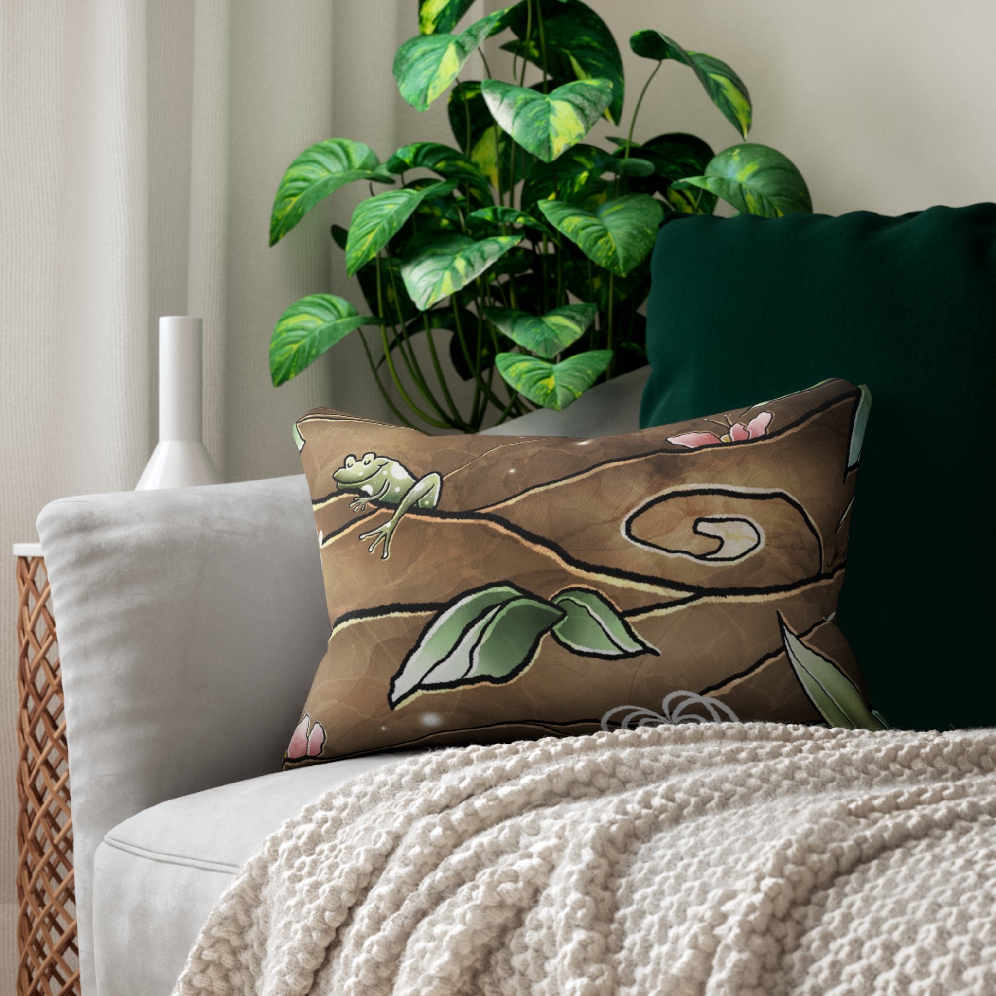 A living room scene with a large variegated green houseplant on a table, a white vase on a side table, a paper bag pillow with a frog illustration, and a beige textured blanket on a sofa with a dark green cushion.