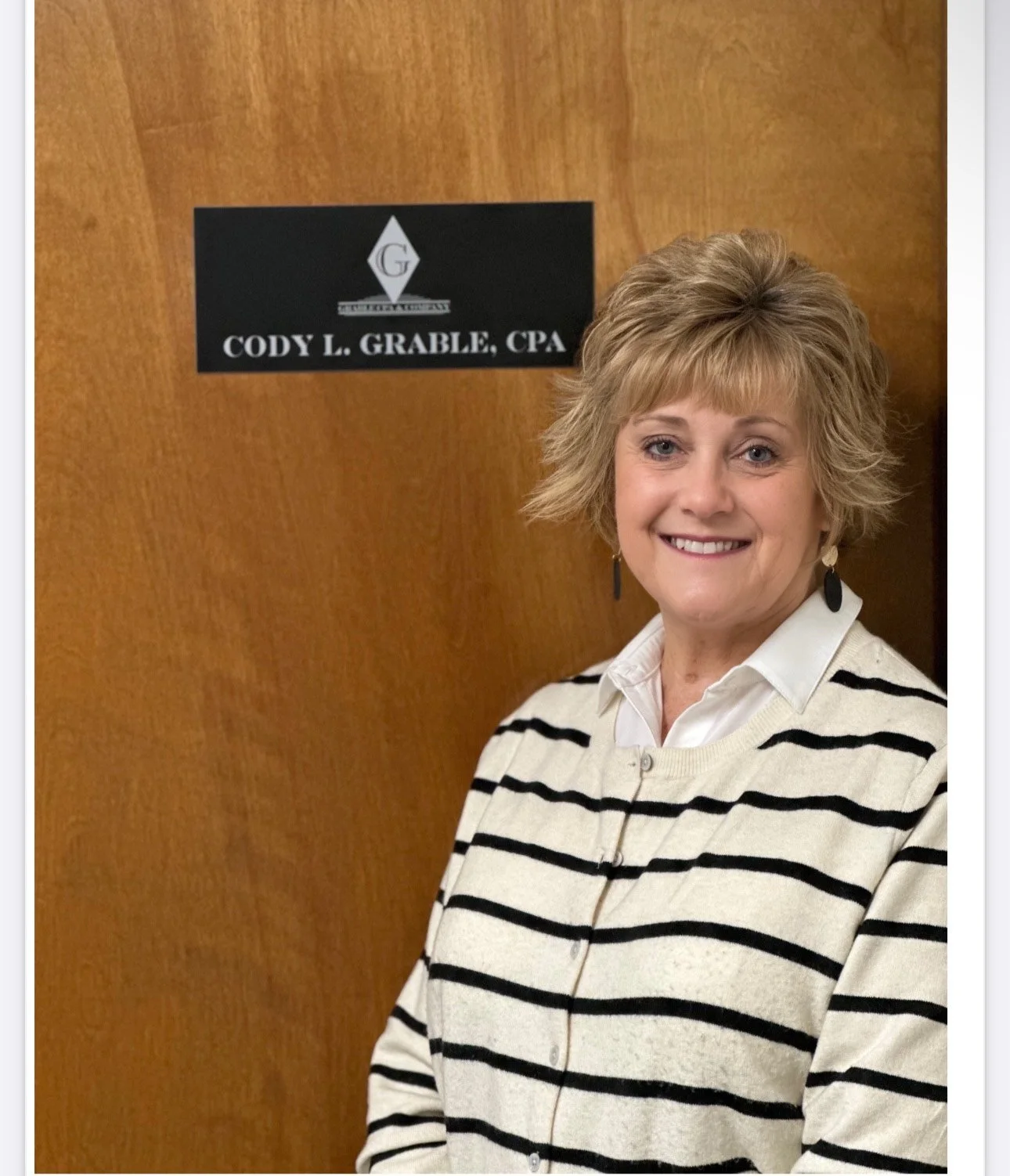 A woman with short blonde hair, wearing a white blouse and a beige cardigan with black stripes, smiling and standing in front of a wooden door with a sign that reads 'Cody L. Grable, CPA'.