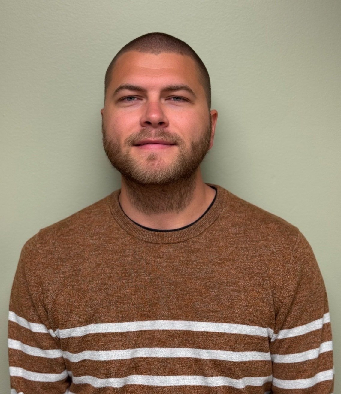 A young man with a short haircut and beard, wearing a brown sweater with white horizontal stripes, standing against a light green wall.