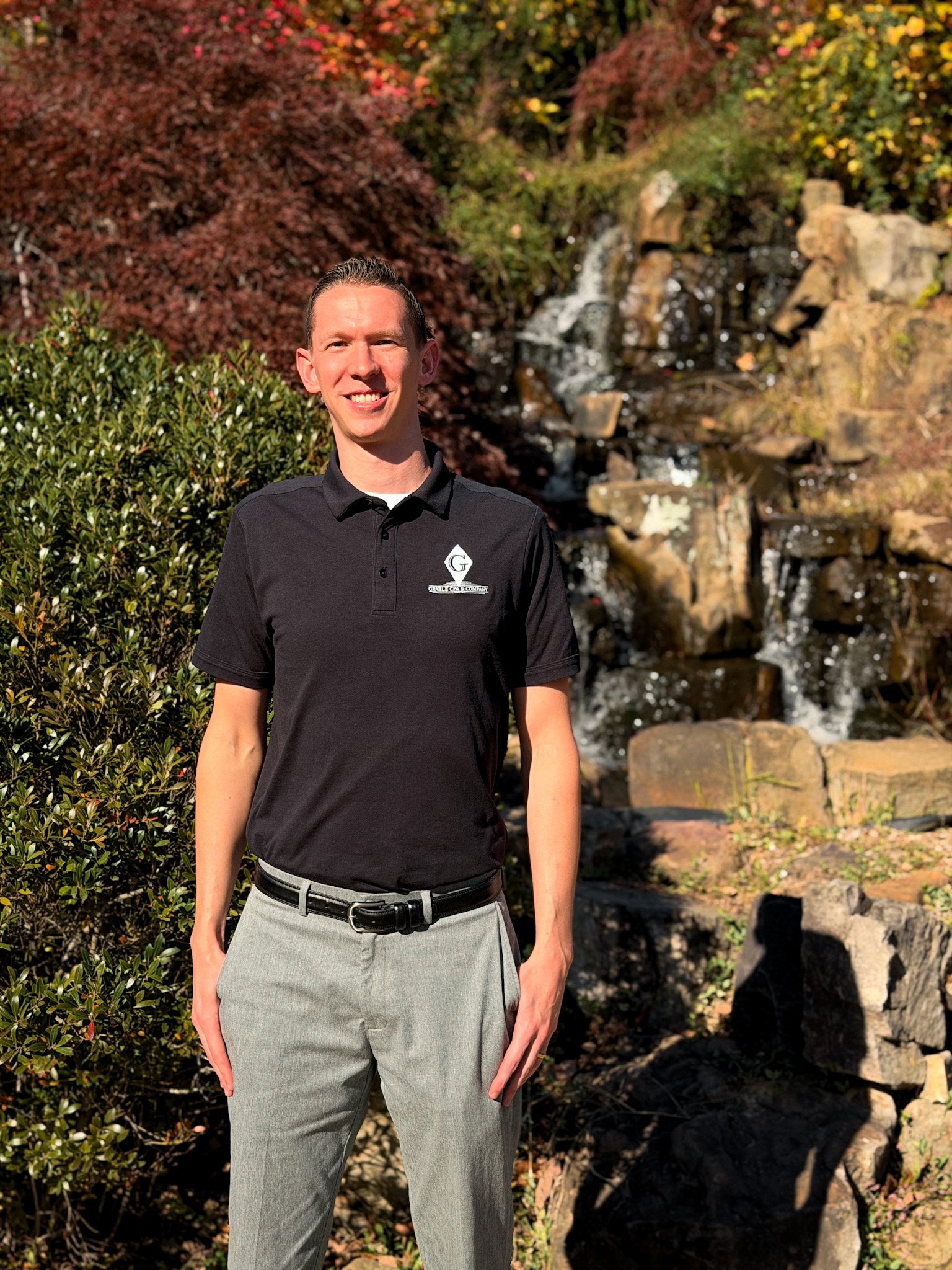 A young man with short brown hair wearing a dark suit, white shirt, and light blue tie, smiling and standing against a plain beige background.