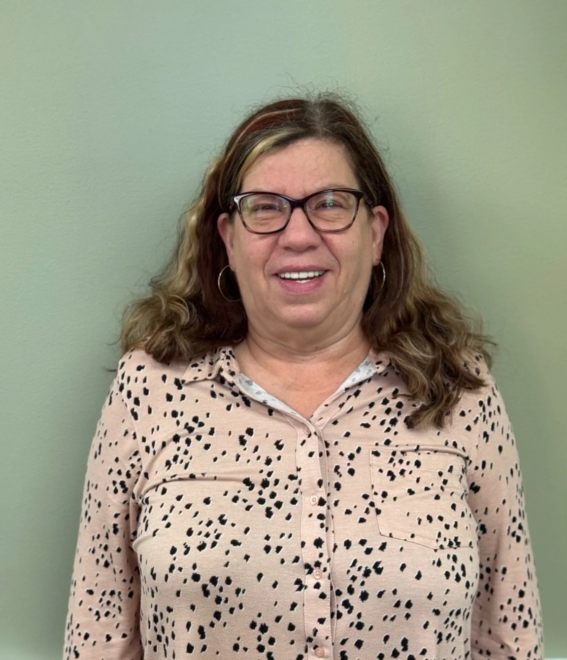 A woman with shoulder-length brown hair, glasses, and hoop earrings, wearing a beige shirt with black spots, smiling and standing against a light green wall.