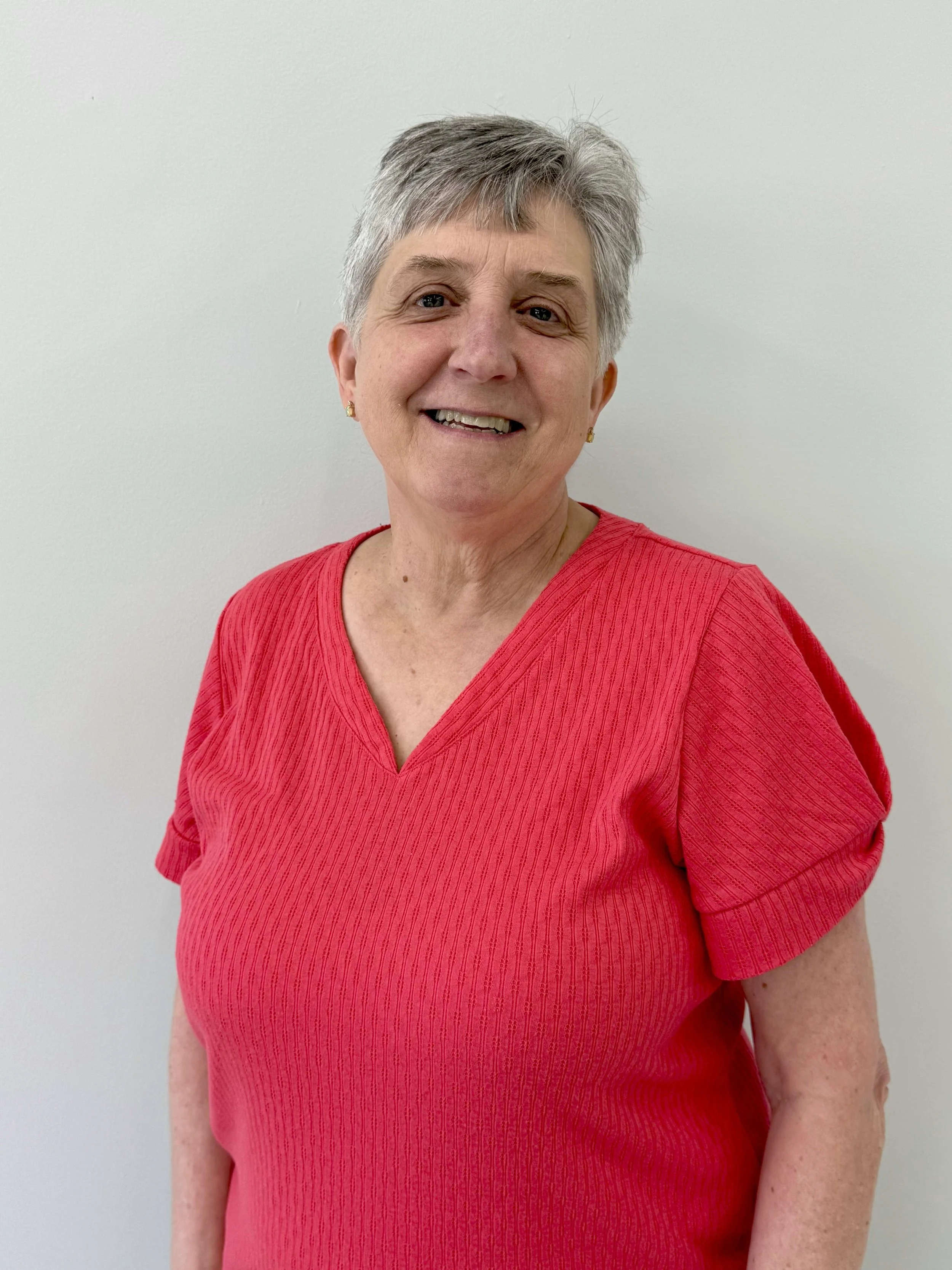 A smiling older woman with short gray hair wearing a red top standing against a plain white wall.