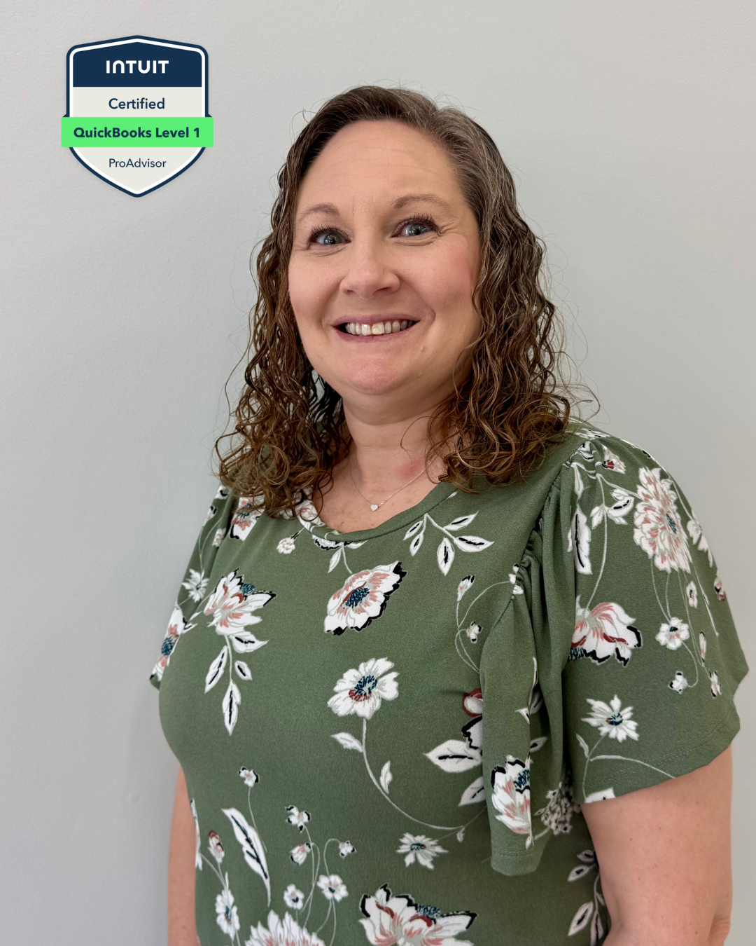 A smiling woman with curly brown hair wearing a green floral dress standing against a plain light gray background.
