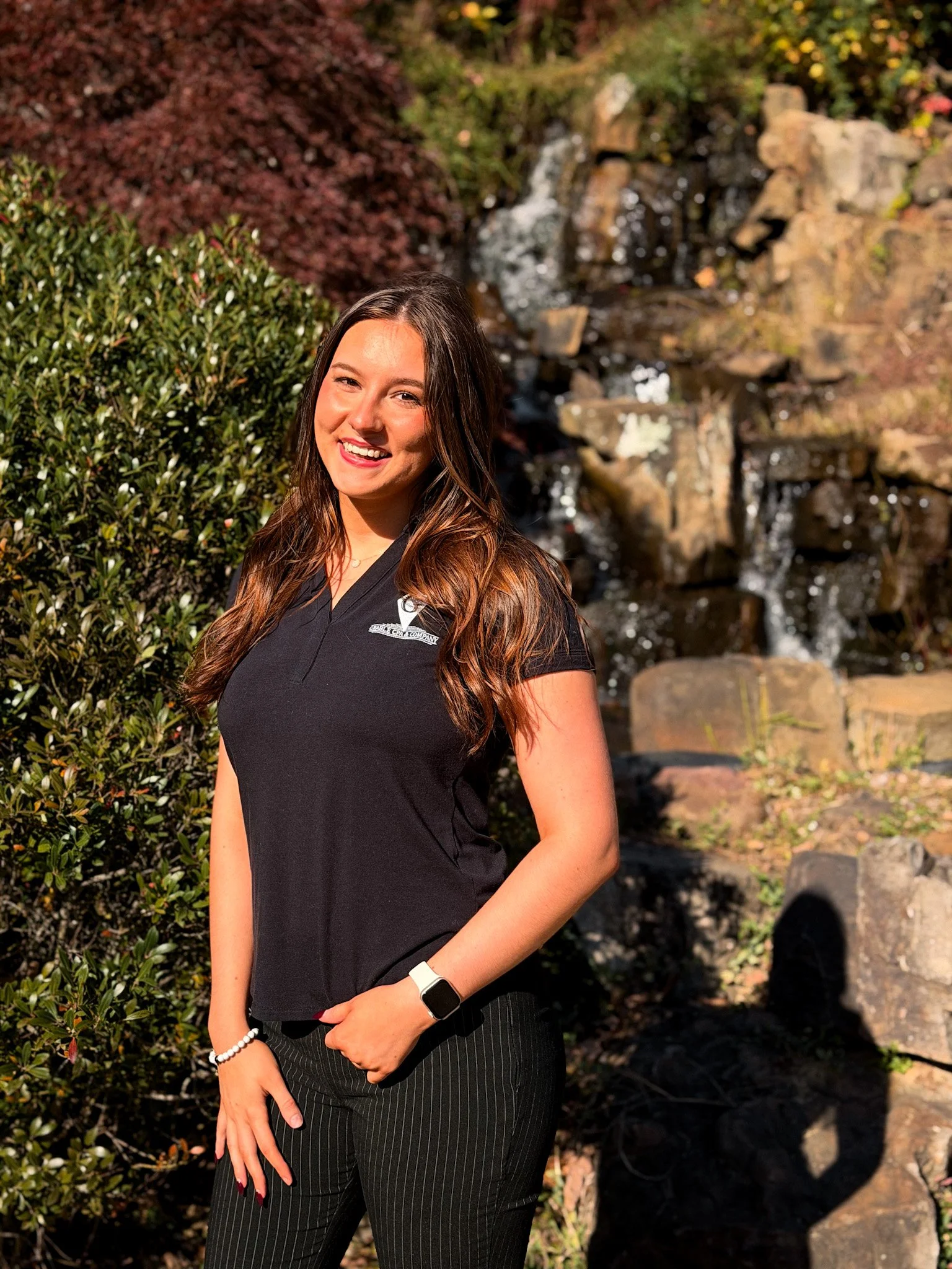 A woman with brown, wavy hair wearing a white cable-knit cardigan and a necklace with a heart-shaped pendant, standing against a plain beige wall.