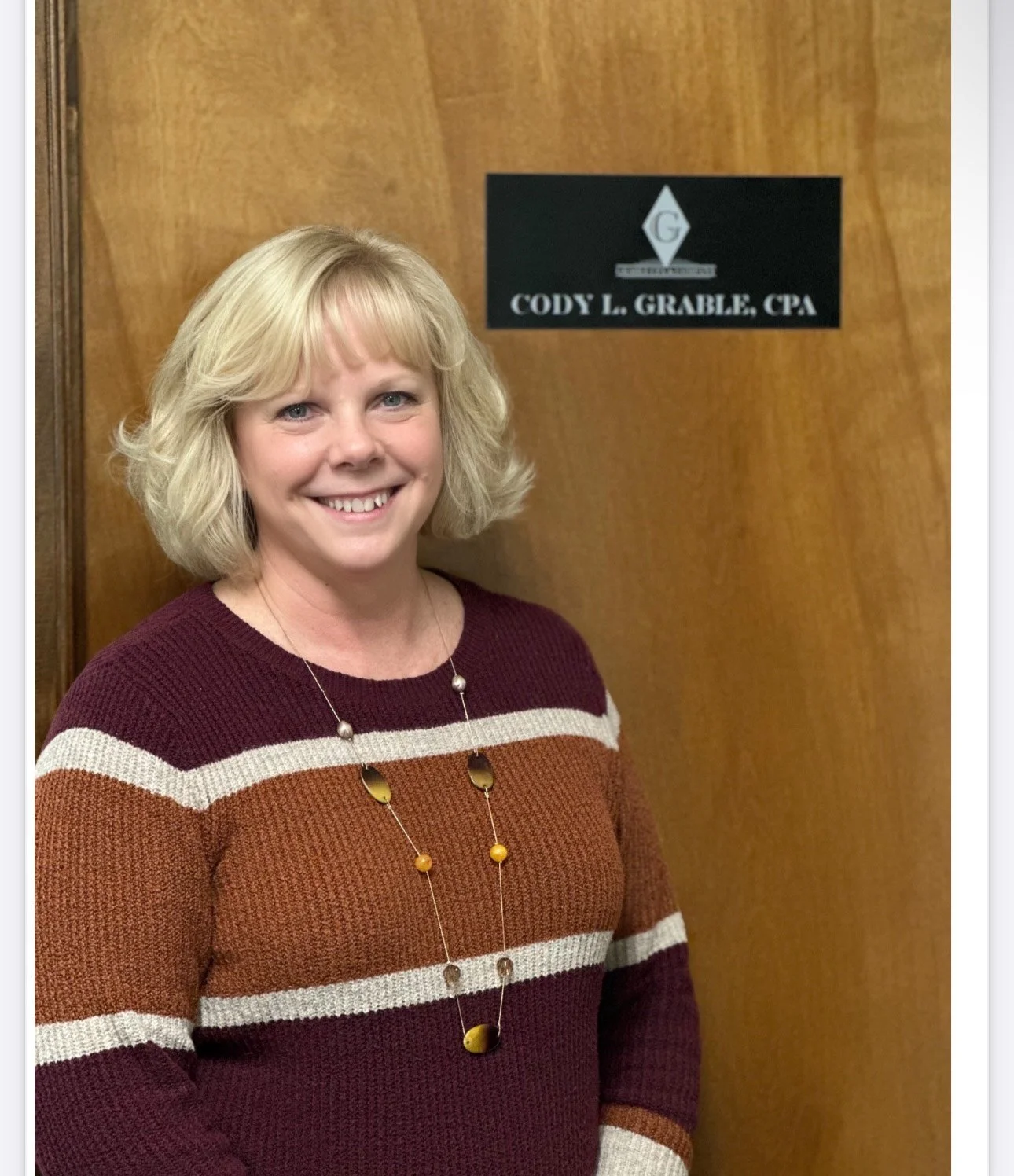 A woman with blonde hair smiling, standing in front of a wooden door, wearing a striped sweater and a long necklace. There is a sign on the door that reads 'Cody L. Grable, CPA'.