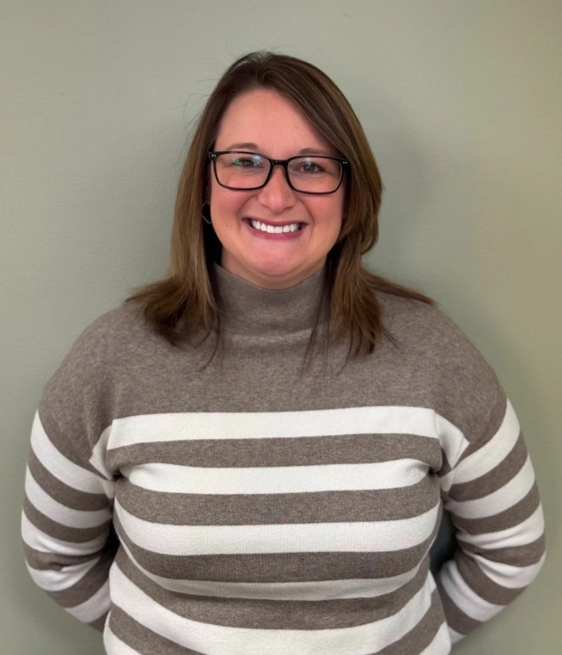 Woman with brown hair, glasses, and a striped sweater smiling against a neutral wall.