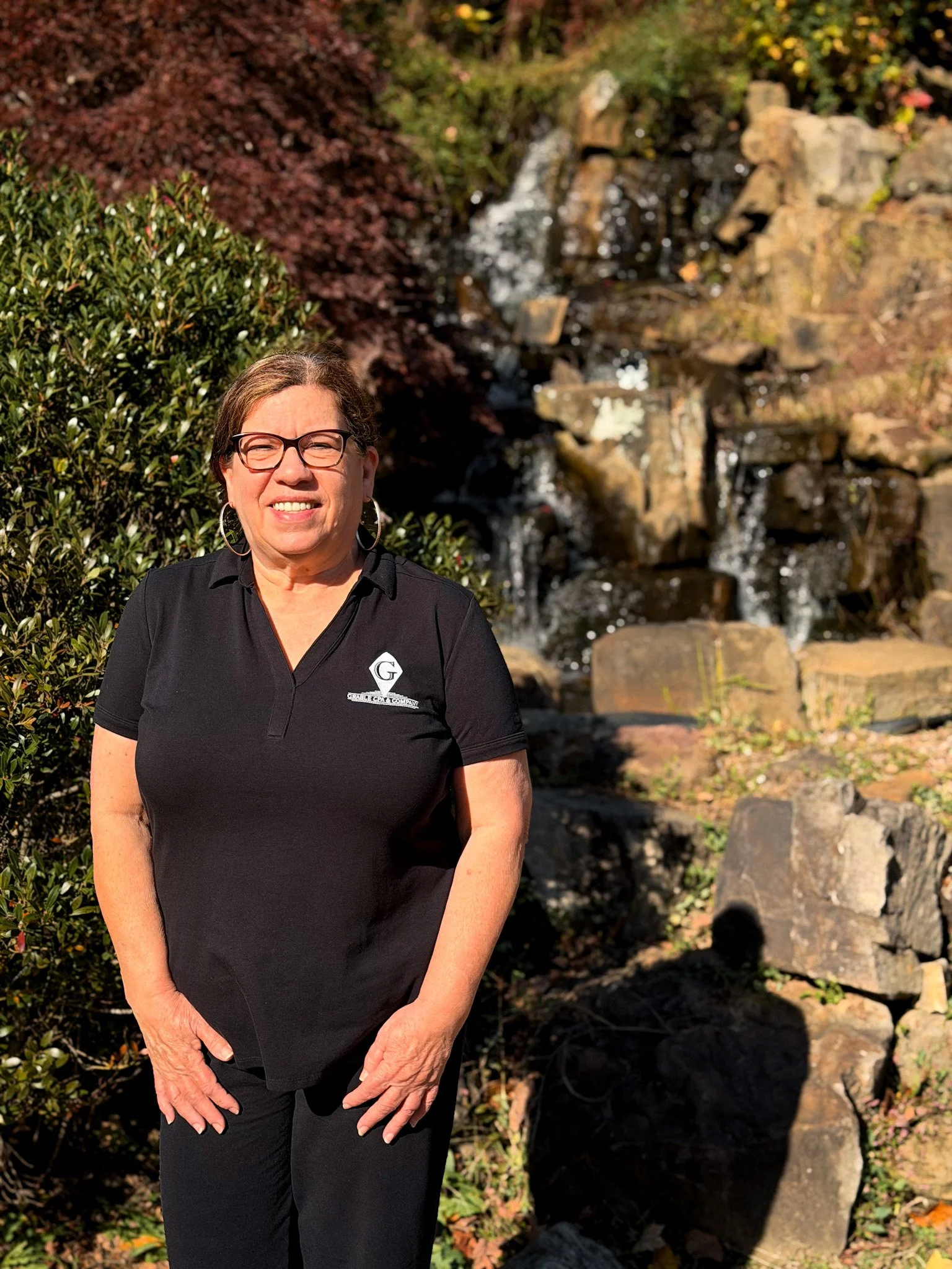 A woman with shoulder-length brown hair, glasses, and hoop earrings, wearing a beige shirt with black spots, smiling and standing against a light green wall.