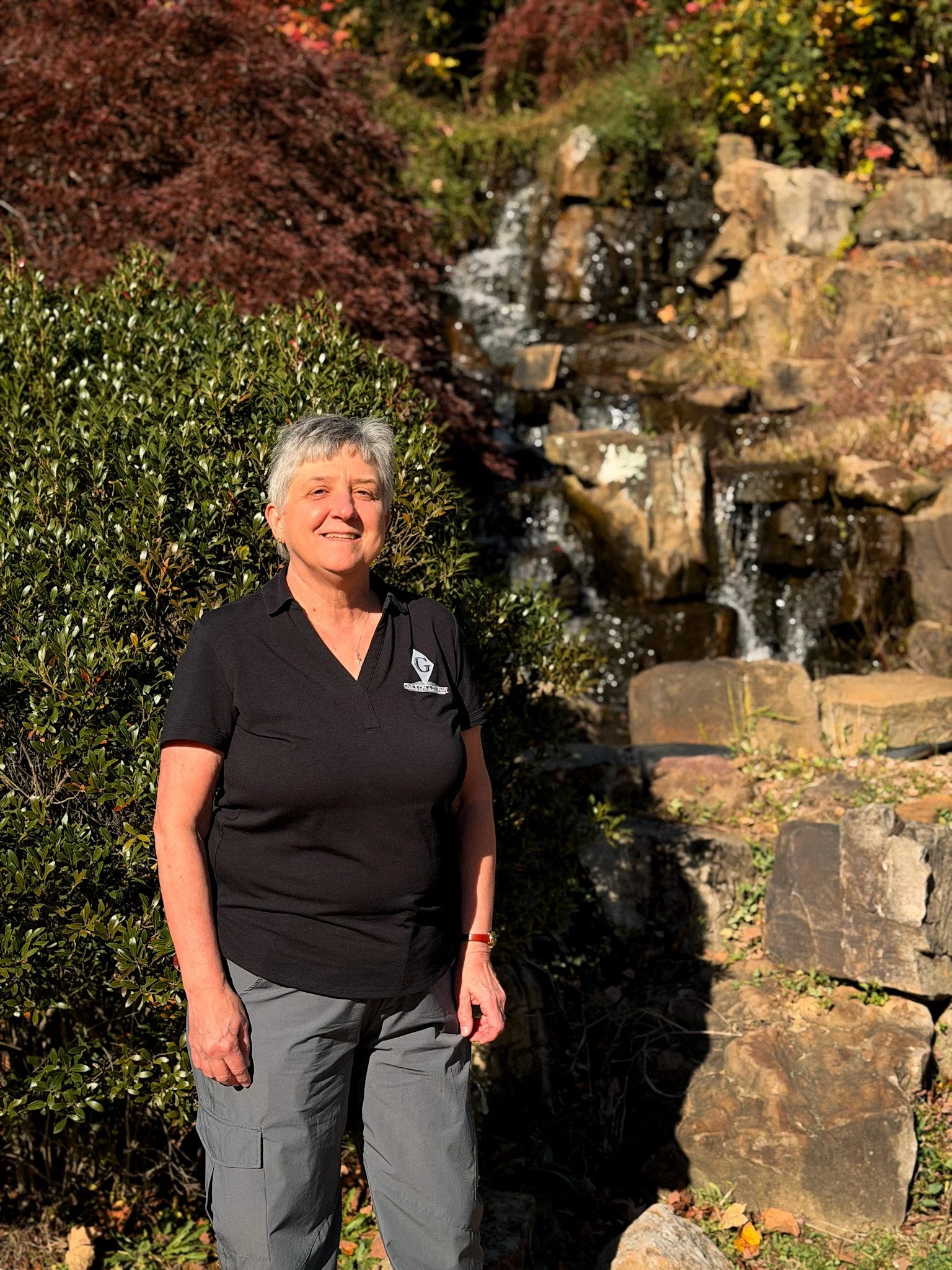 A smiling older woman with short gray hair wearing a red top standing against a plain white wall.