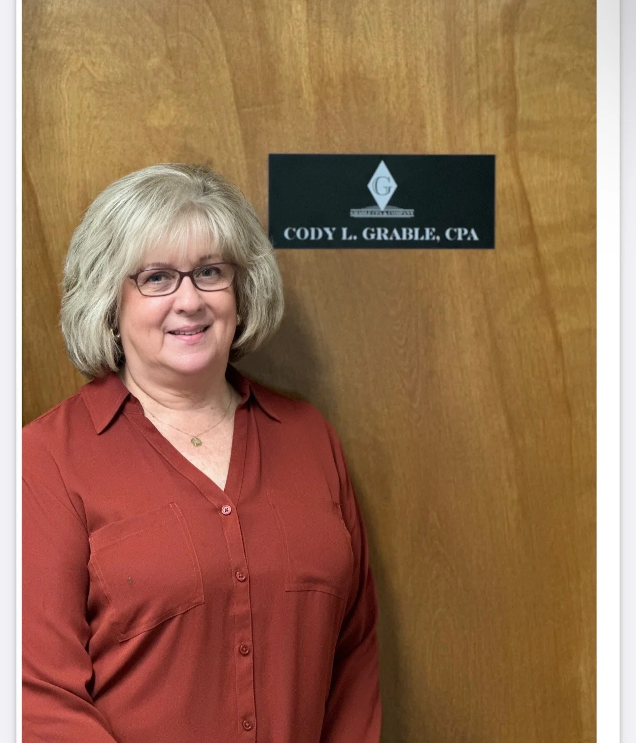 A woman with blonde hair, glasses, and wearing a red button-up shirt standing next to a door with a sign that reads 'Cody L. Grable, CPA'.