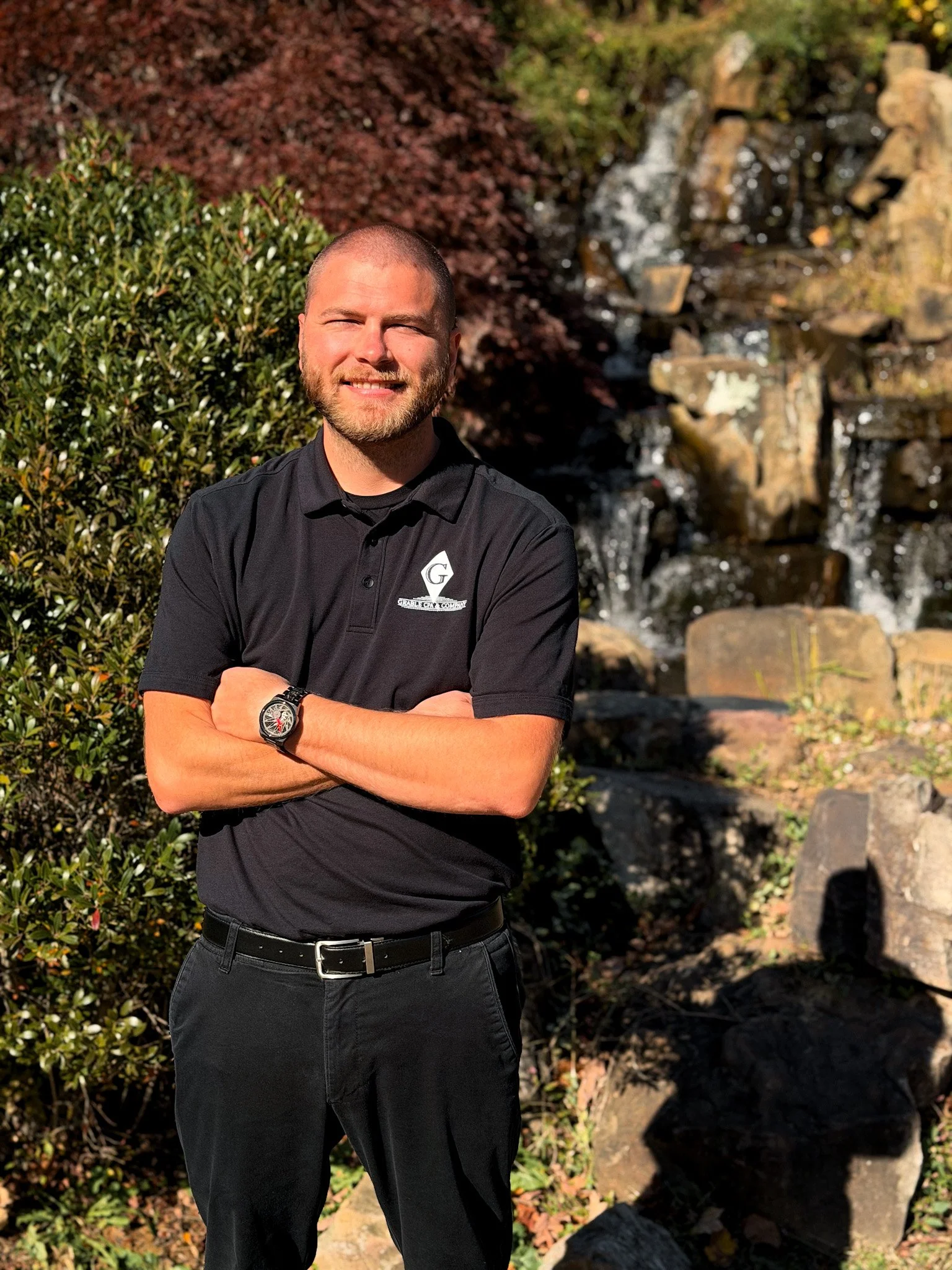 A young man with a short haircut and beard, wearing a brown sweater with white horizontal stripes, standing against a light green wall.