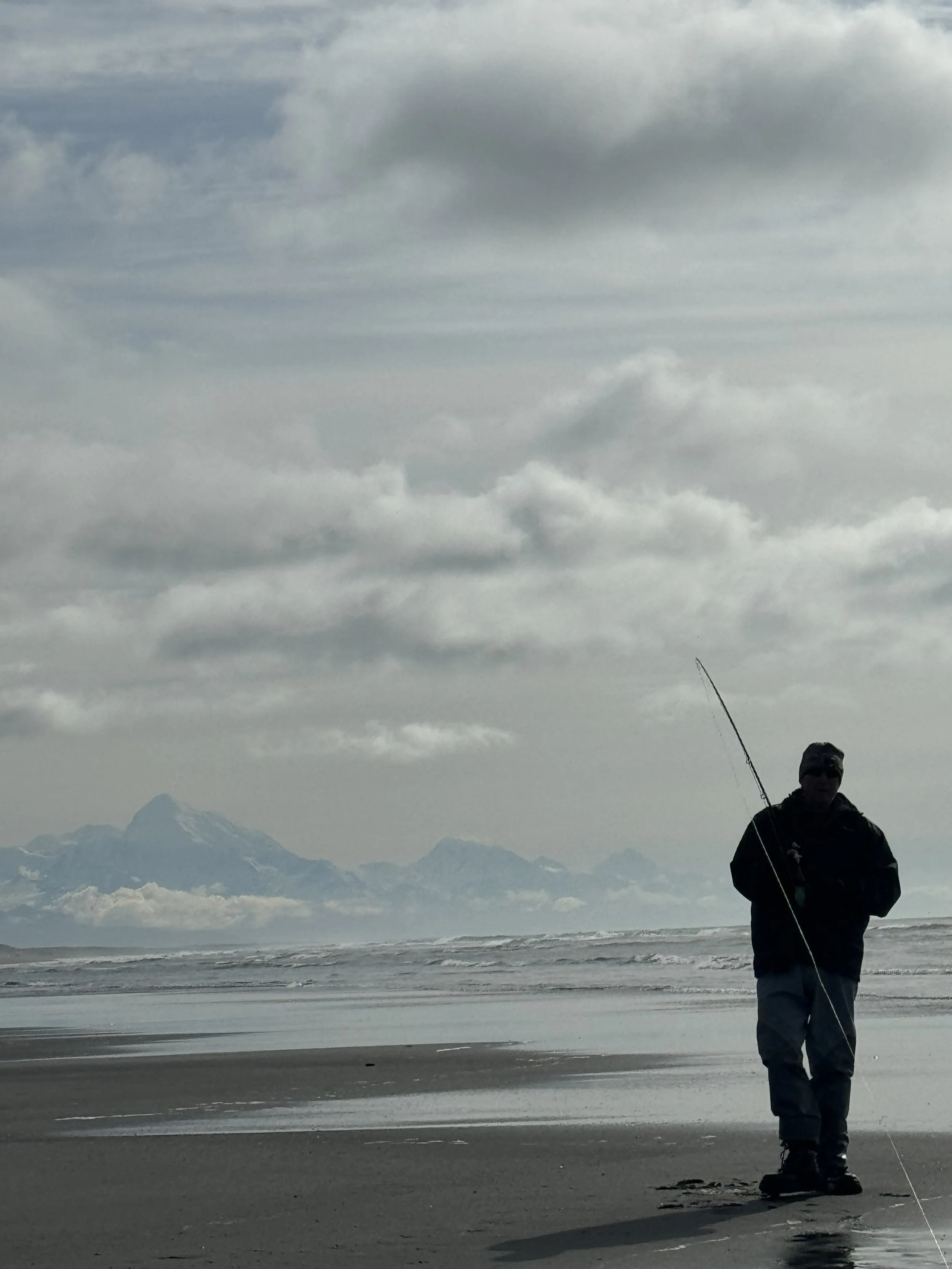 Young boy holding a large fish while kneeling in shallow water on a sunny day with a blue sky and mountains in the background.