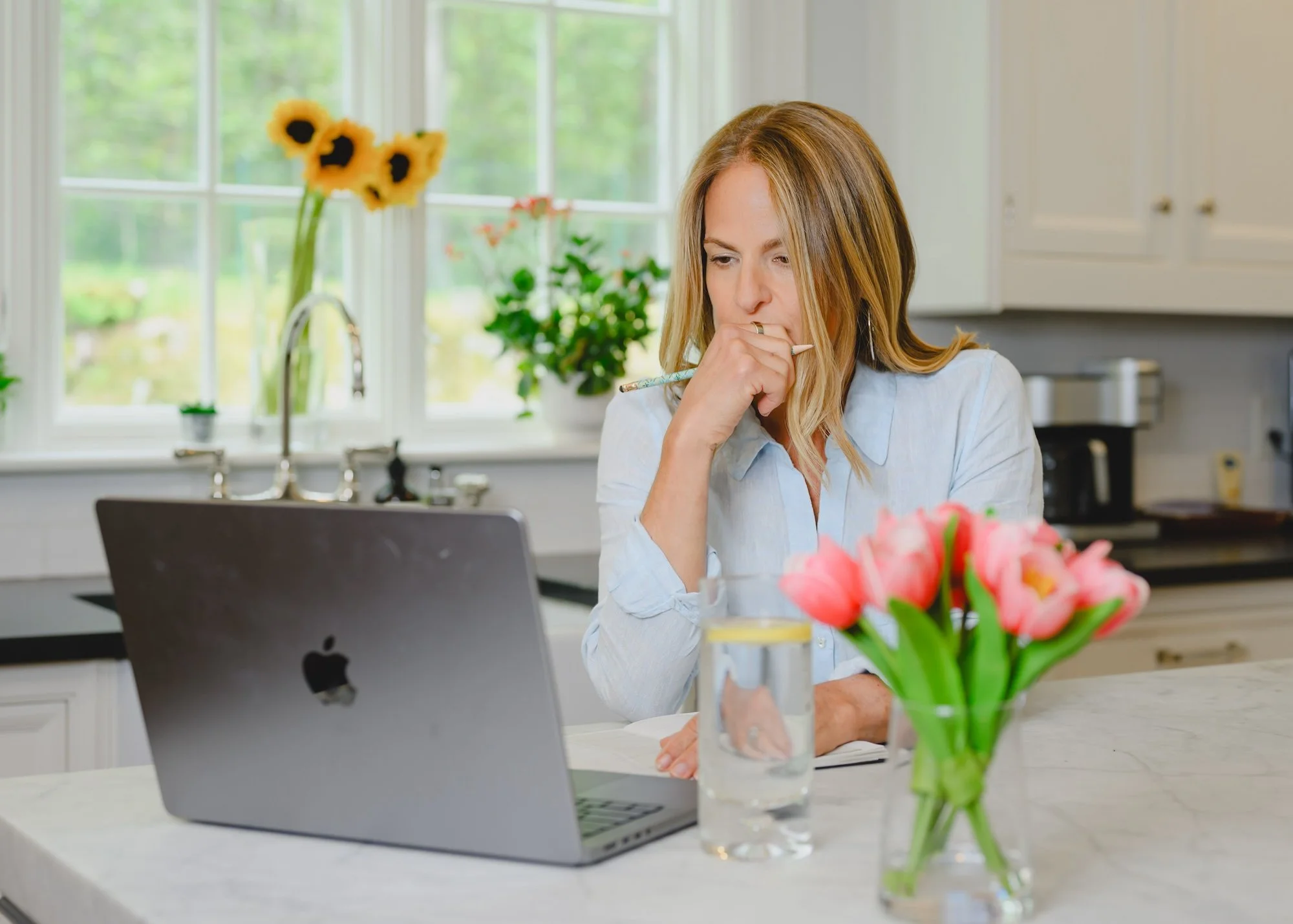 Woman with blonde hair sitting at a white kitchen counter, working or thinking, with a laptop in front of her. The background has a window with natural light, a sink, and kitchen cabinets. There is a bouquet of pink tulips in a glass vase on the counter.