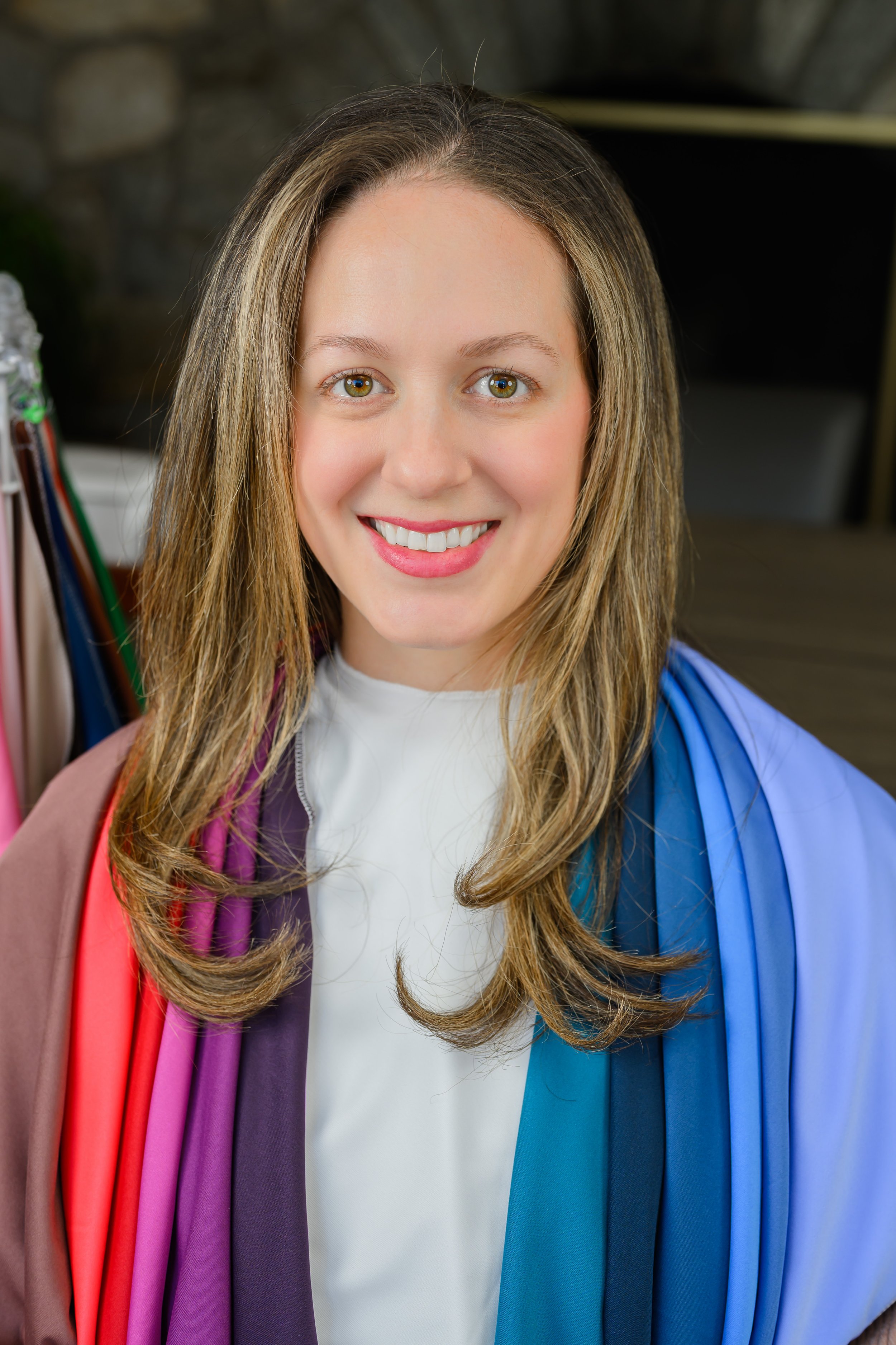 Close-up of a young woman with long light brown hair, green eyes, and a bright smile, wearing a white shirt and a blue graduation gown, carrying colorful stoles.