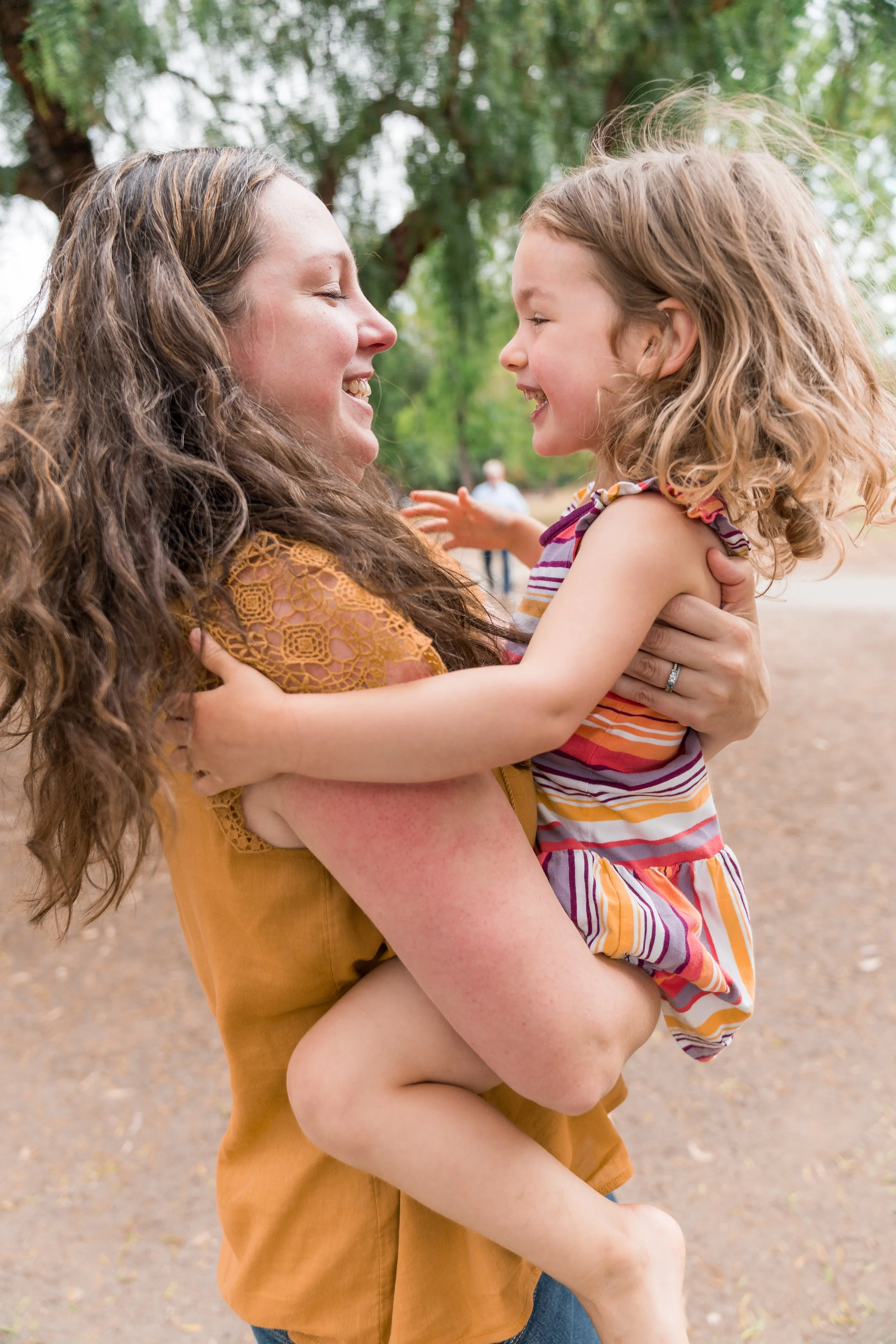 A woman with long, curly brown hair smiling while holding a young girl with curly blonde hair outdoors.