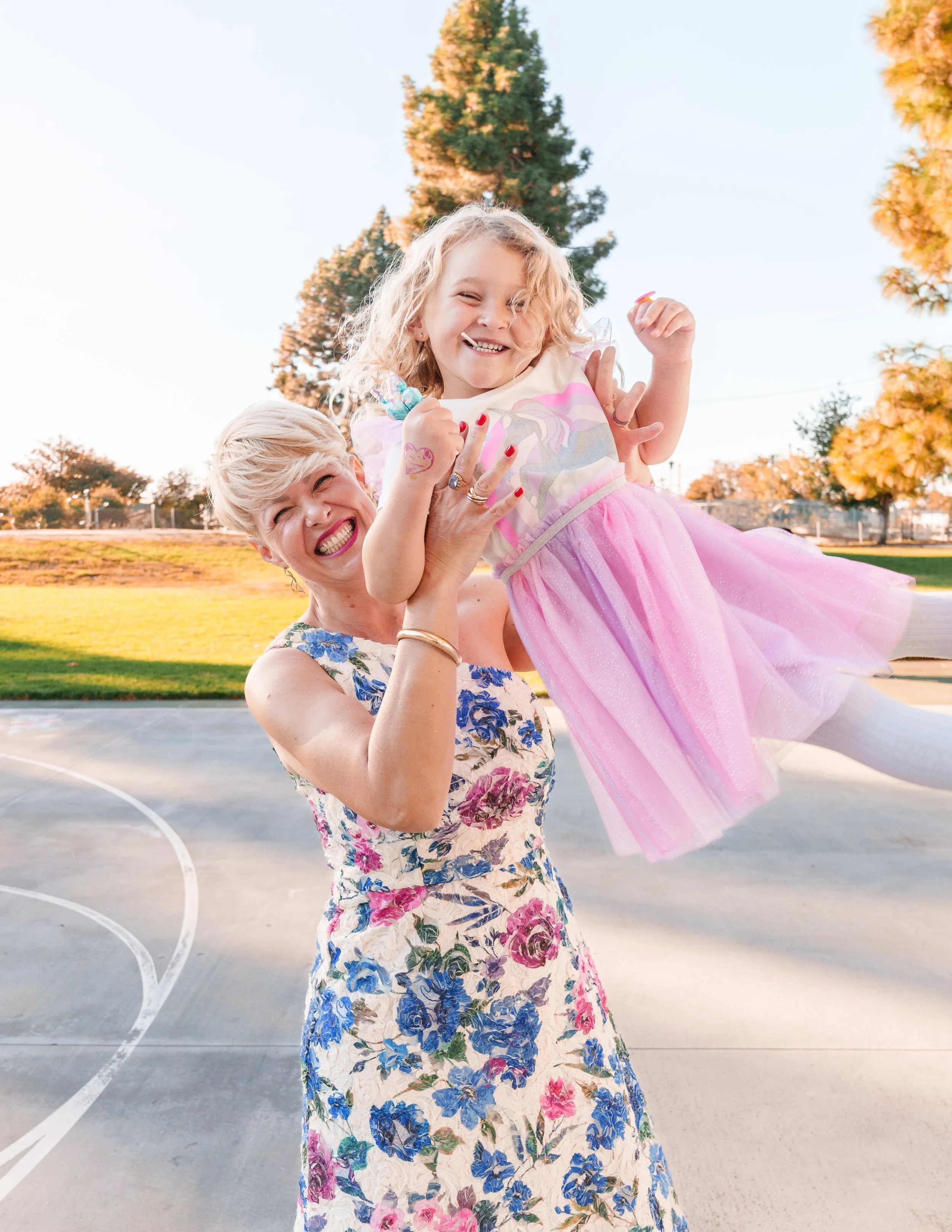 A joyful young girl wearing a pink tutu and a woman in a floral dress lifting her up holding her from underneath on a basketball court at sunset, with trees in the background.