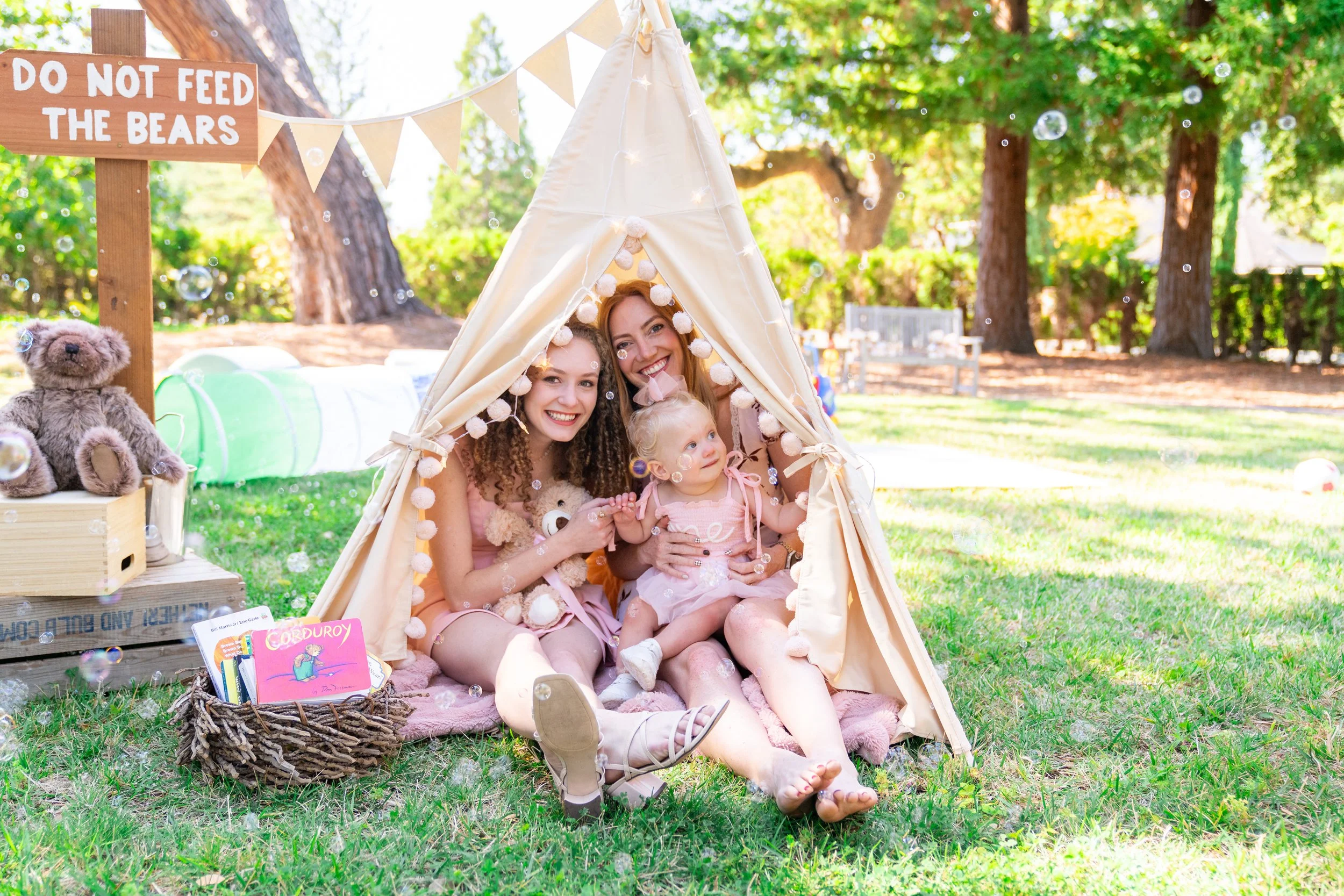 Three women and a young girl sitting inside a cream-colored fabric teepee decorated with pom-poms, outdoors in a park with green trees and sunlight. There are bubbles floating around, a teddy bear, and a small basket with children's books and toys nearby. A wooden sign reads 'Do not feed the bears'.