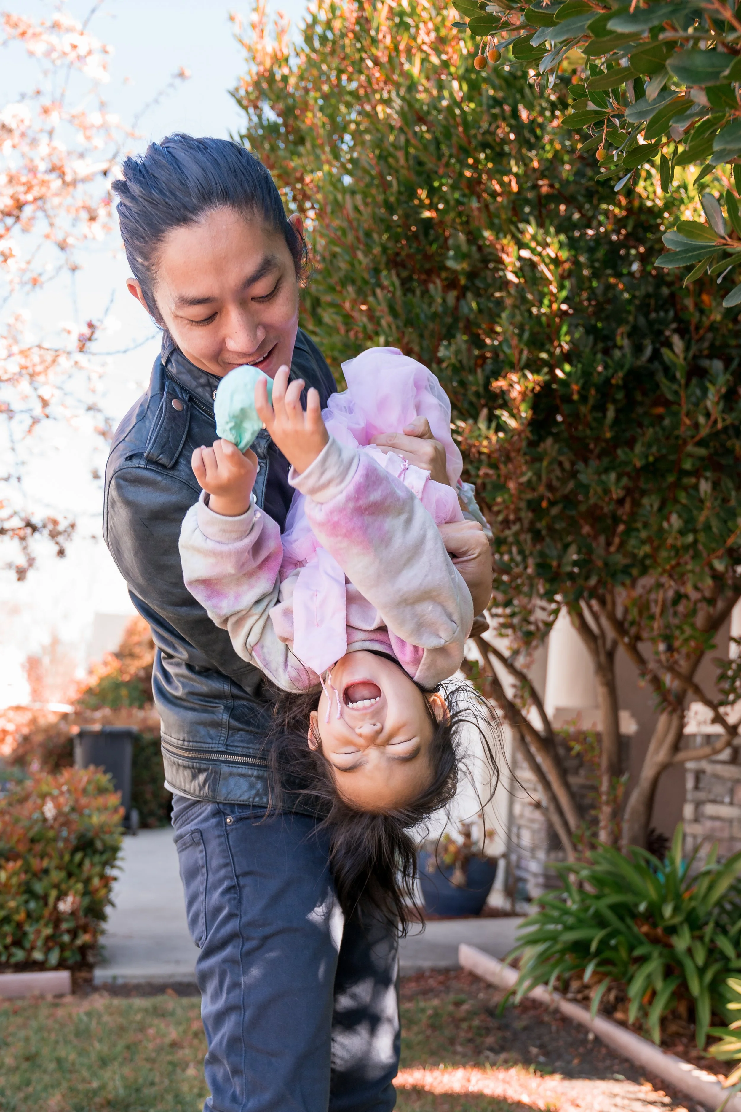 A man with long dark hair wearing a black leather jacket and dark jeans, holding a young girl upside down, both smiling and laughing outside in a garden or backyard with bushes and trees with autumn leaves.