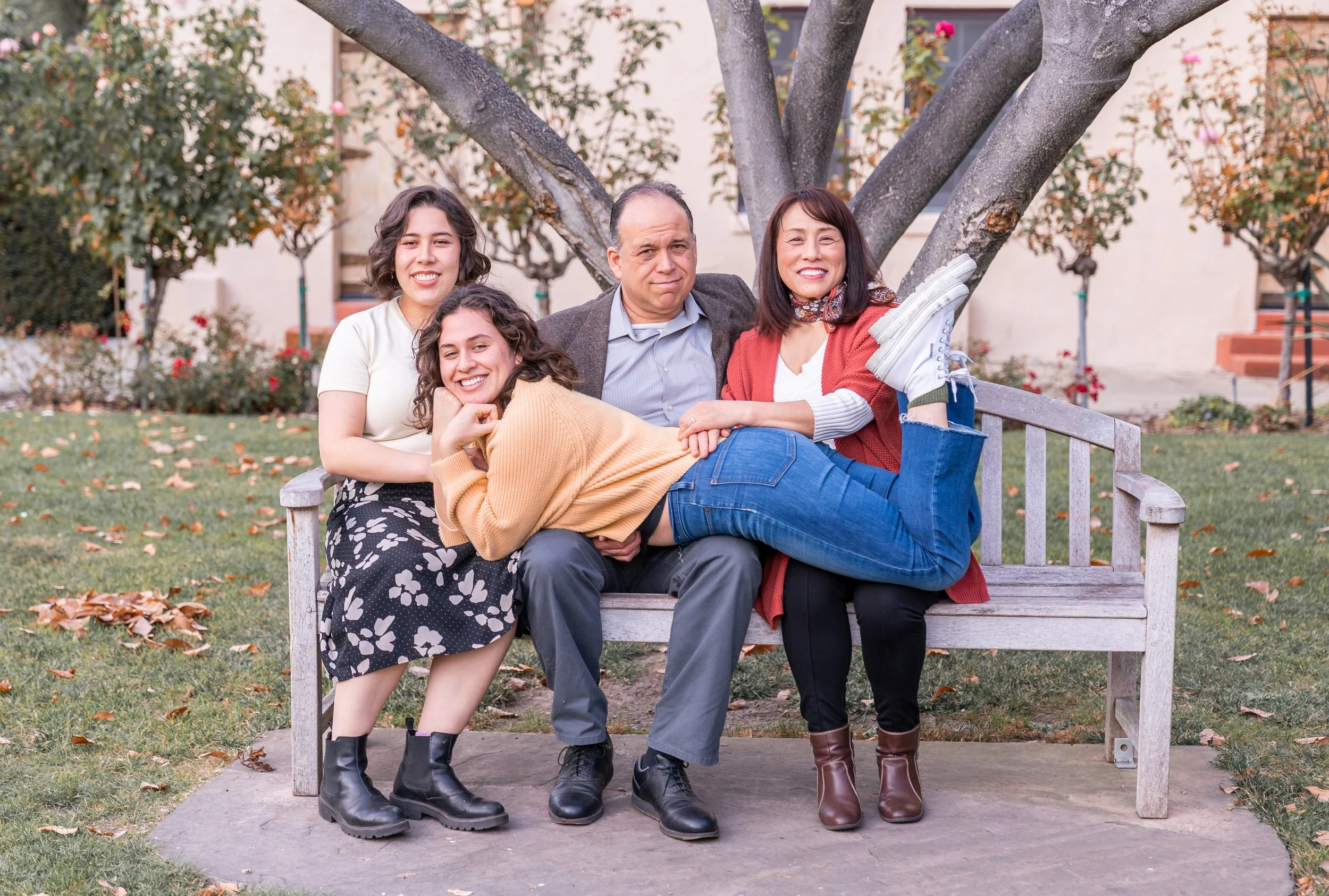 Family of five sitting on a park bench with a large tree and fallen leaves in the background.