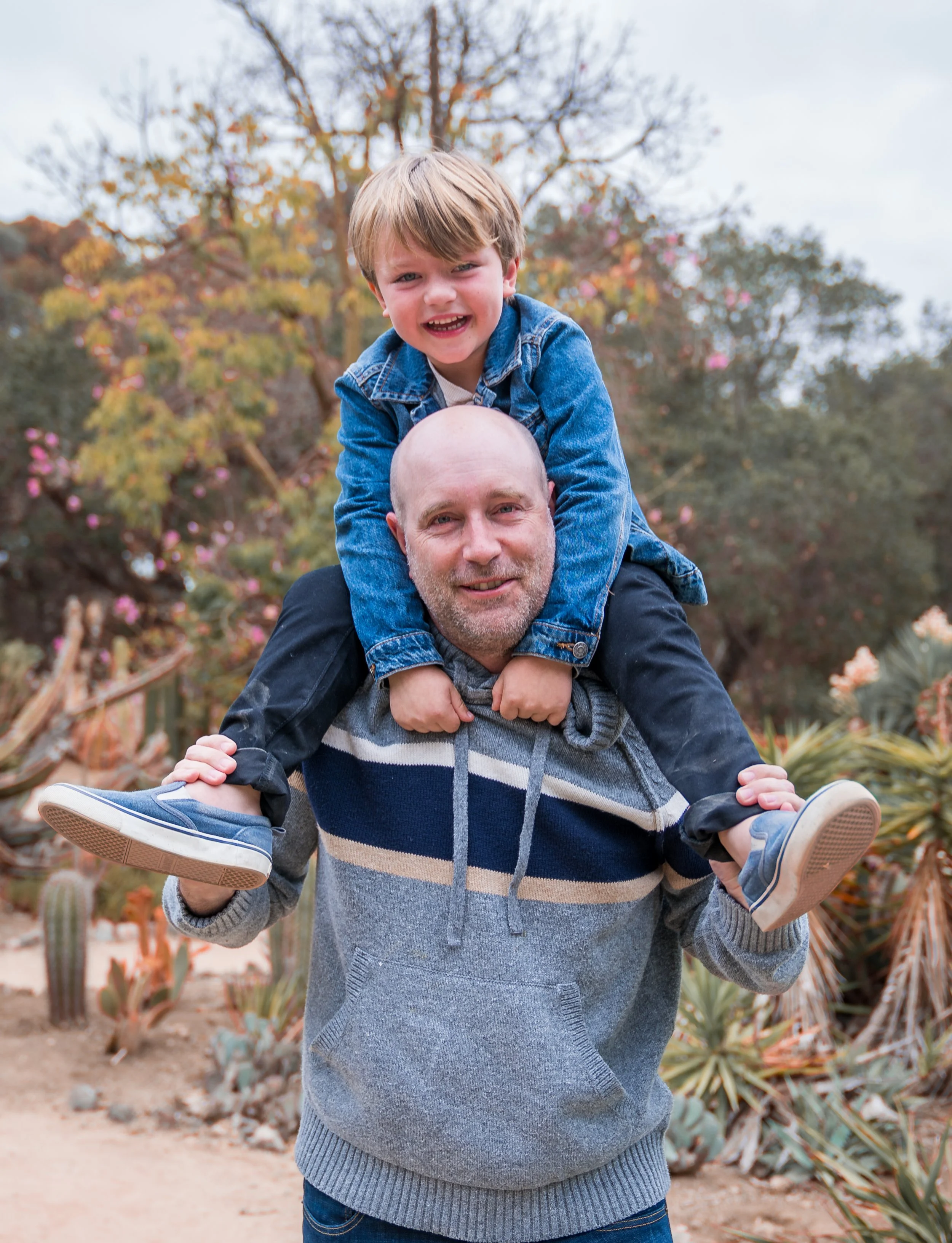 A man giving a child a piggyback ride outdoors, surrounded by plants and trees with autumn foliage.