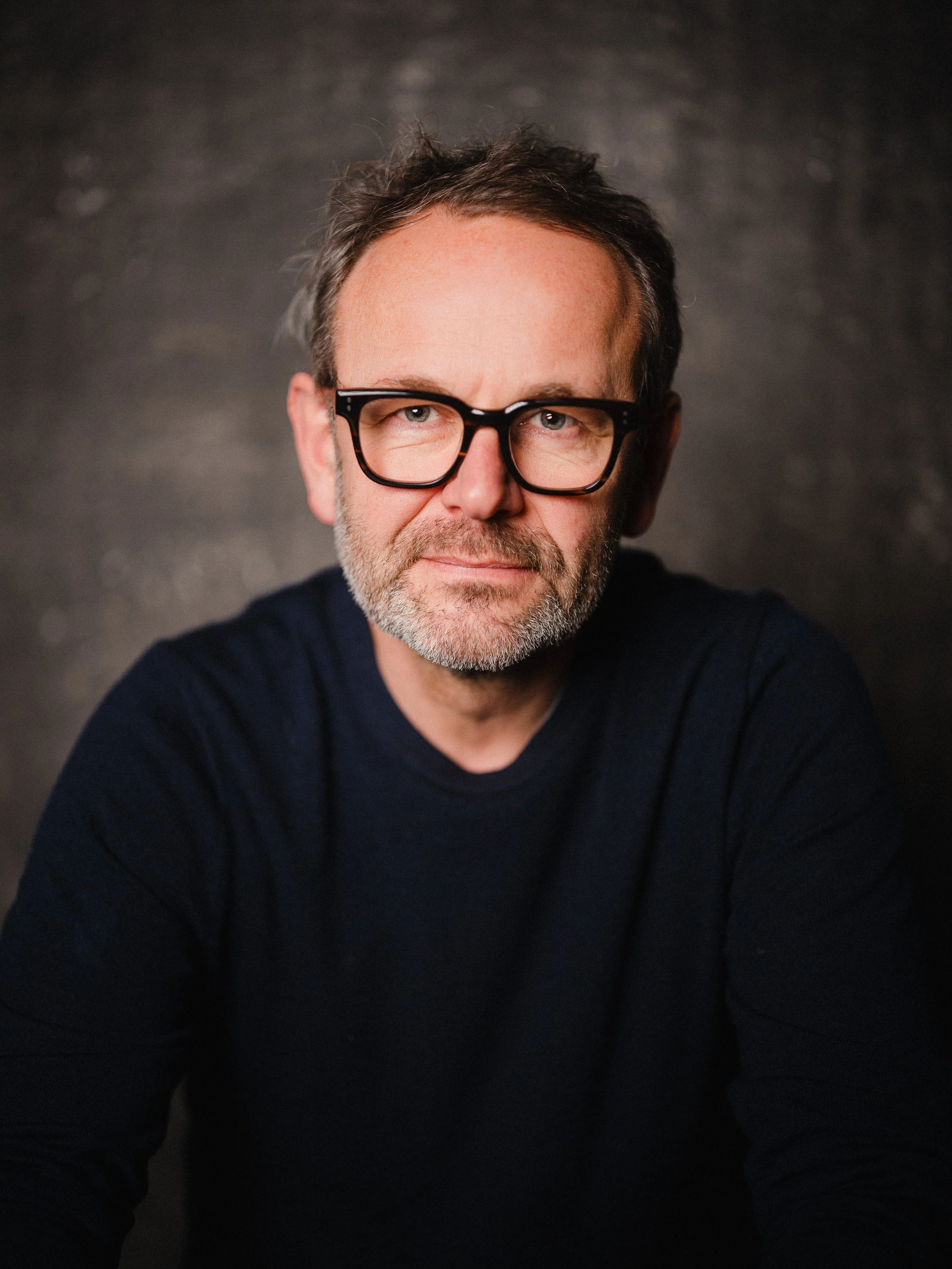 Porträt eines Mannes mit Brille, grauem Bart, dunkelblauem Pullover, vor dunklem Hintergrund.