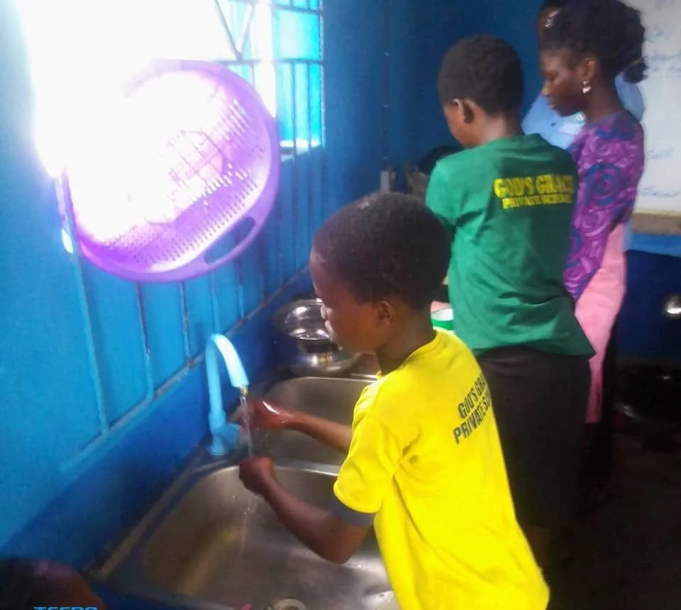 Children washing dishes together in a kitchen with blue walls, a purple fan, and a window.