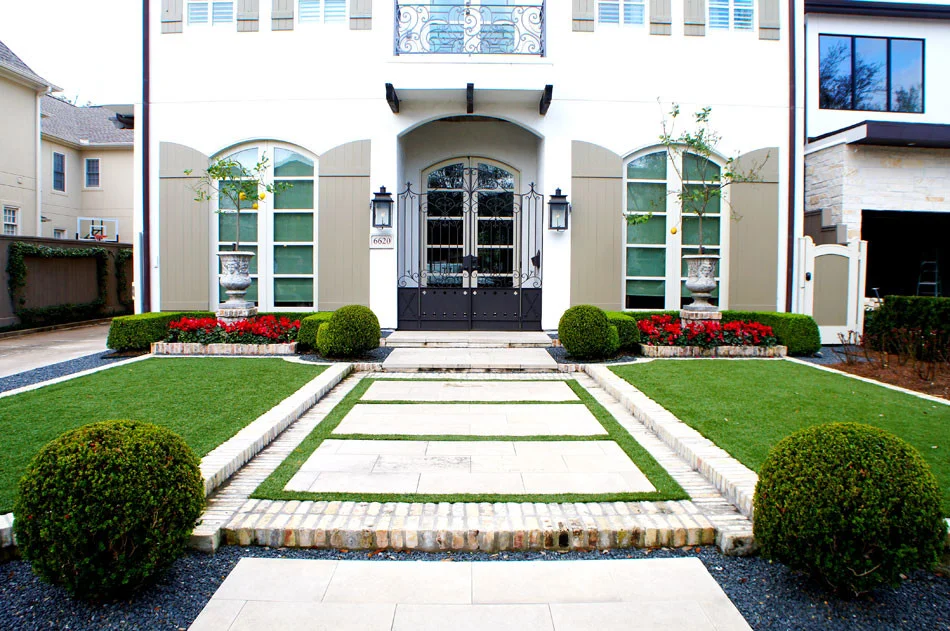 Front view of this French Contemporary Villa captures the symmetry of the front lawns and walking path. Sharply trimmed boxwood hedges frame color beds of Cyclamen which accent mirrored sculpted planters. White brick separates the artificial lawn from borders of blackstar gravel.