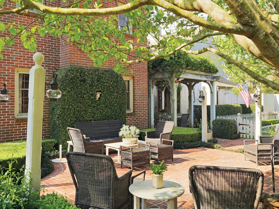 Red brick patio with woven outdoor seating. Ivy covered walls and wisteria covered pergola that frames outdoor dining area. Lamp posts with lanterns and white picket fence add extra detail to this New England style home in Houston, TX.