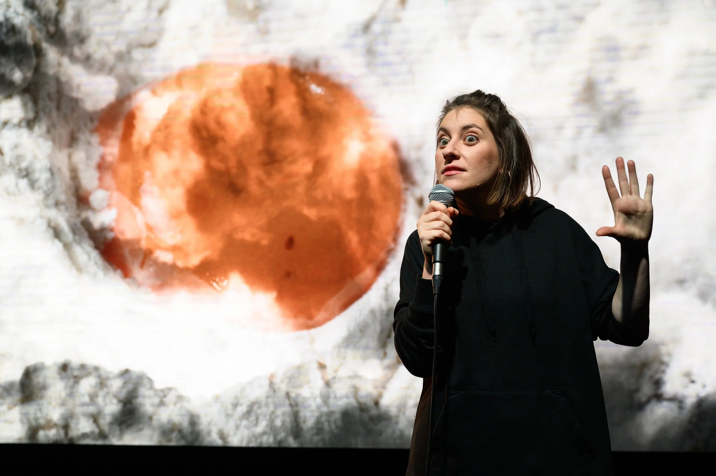 Mulher com cabelo curto segurando um microfone na frente de uma projeção de uma explosão na lua, vestindo uma blusa preta, aparentando estar falando ou apresentando.