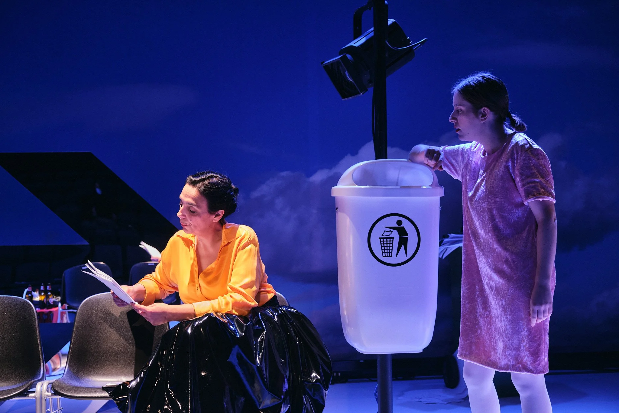 Duas mulheres em cena de teatro, uma sentada lendo um roteiro e outra de pé colocando lixo na lixeira, com iluminação azulada, ao fundo um piano e uma garrafa de bebidas.
