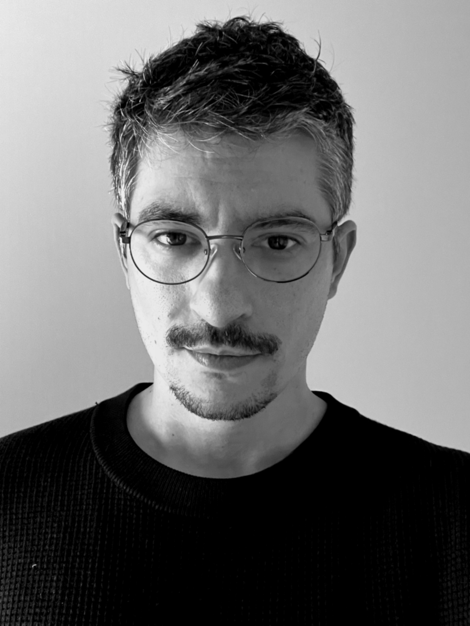 Retrato de um homem com cabelo castanho escuro, usando óculos de aros redondos e uma blusa preta, olhando para a câmera, em preto e branco.