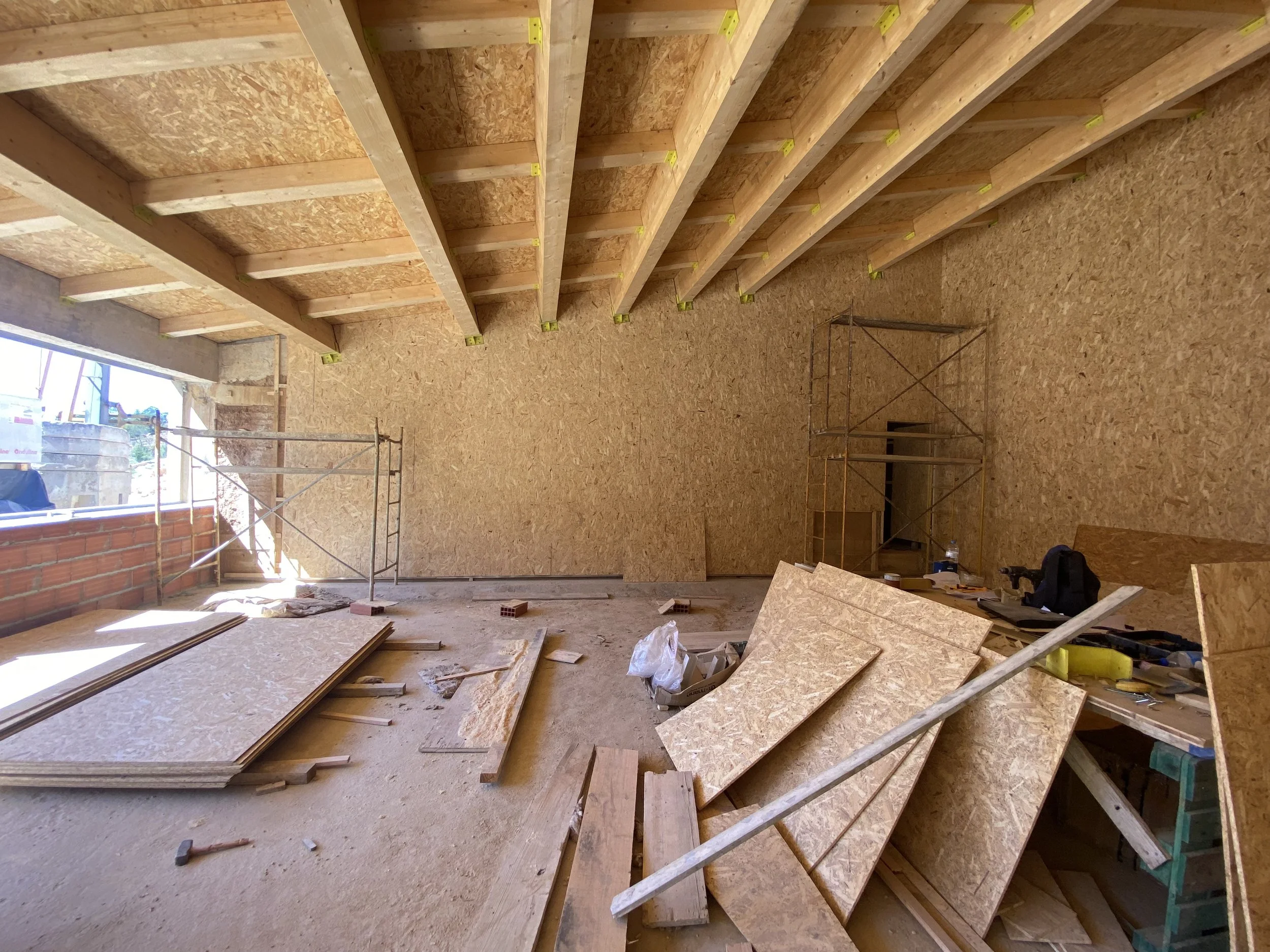 Interior de um cômodo em construção com estrutura de madeira no teto e paredes de OSB, ferramentas e materiais de construção espalhados pelo chão.