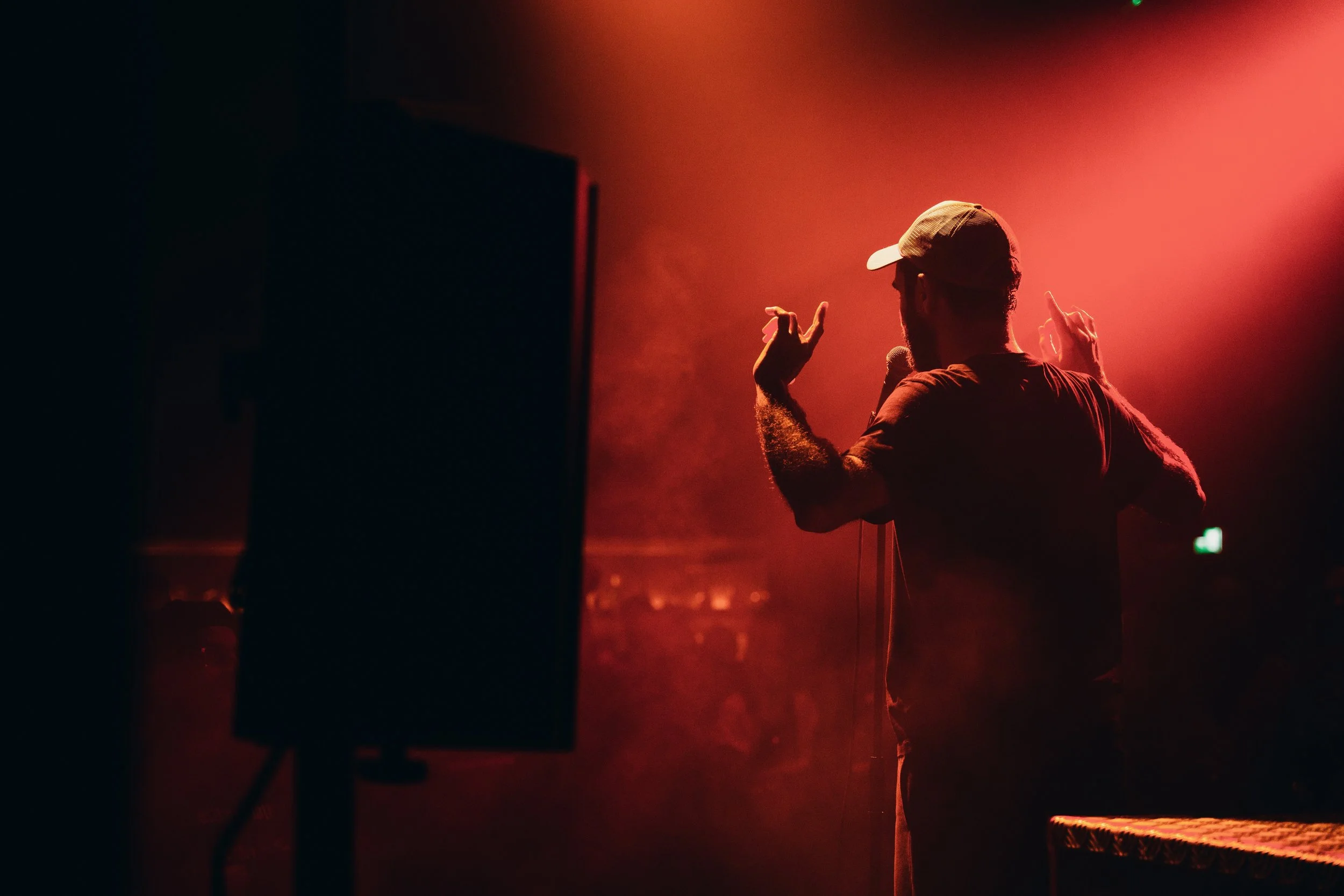 Homem com boné fazendo um discurso ou apresentação em um palco com iluminação vermelha.