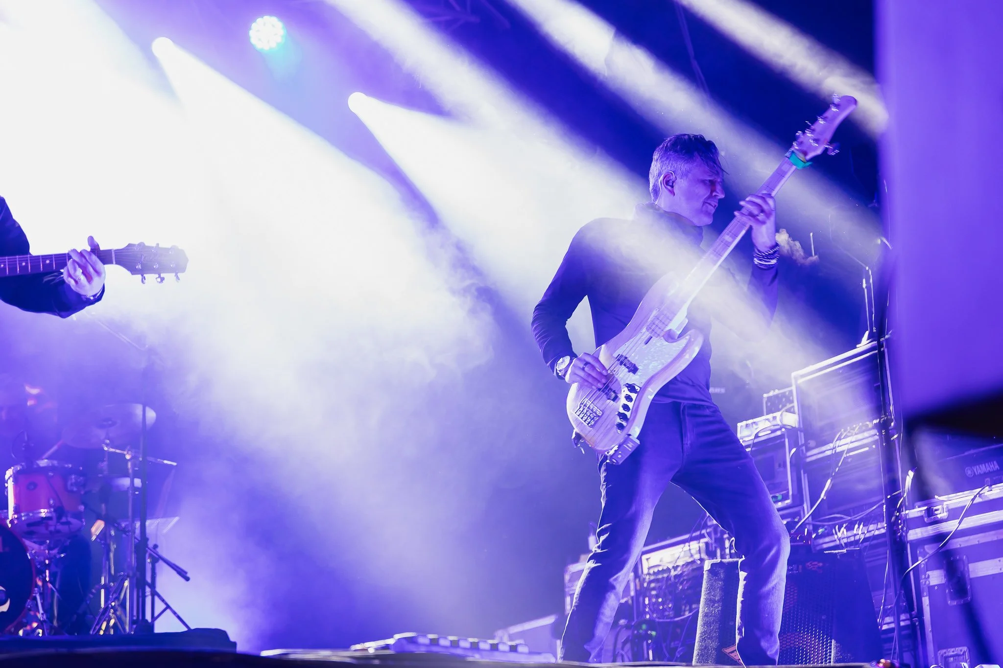 Músico tocando guitarra elétrica em um palco iluminado com luzes azuis e brancas, acompanhado de outros instrumentos musicais.