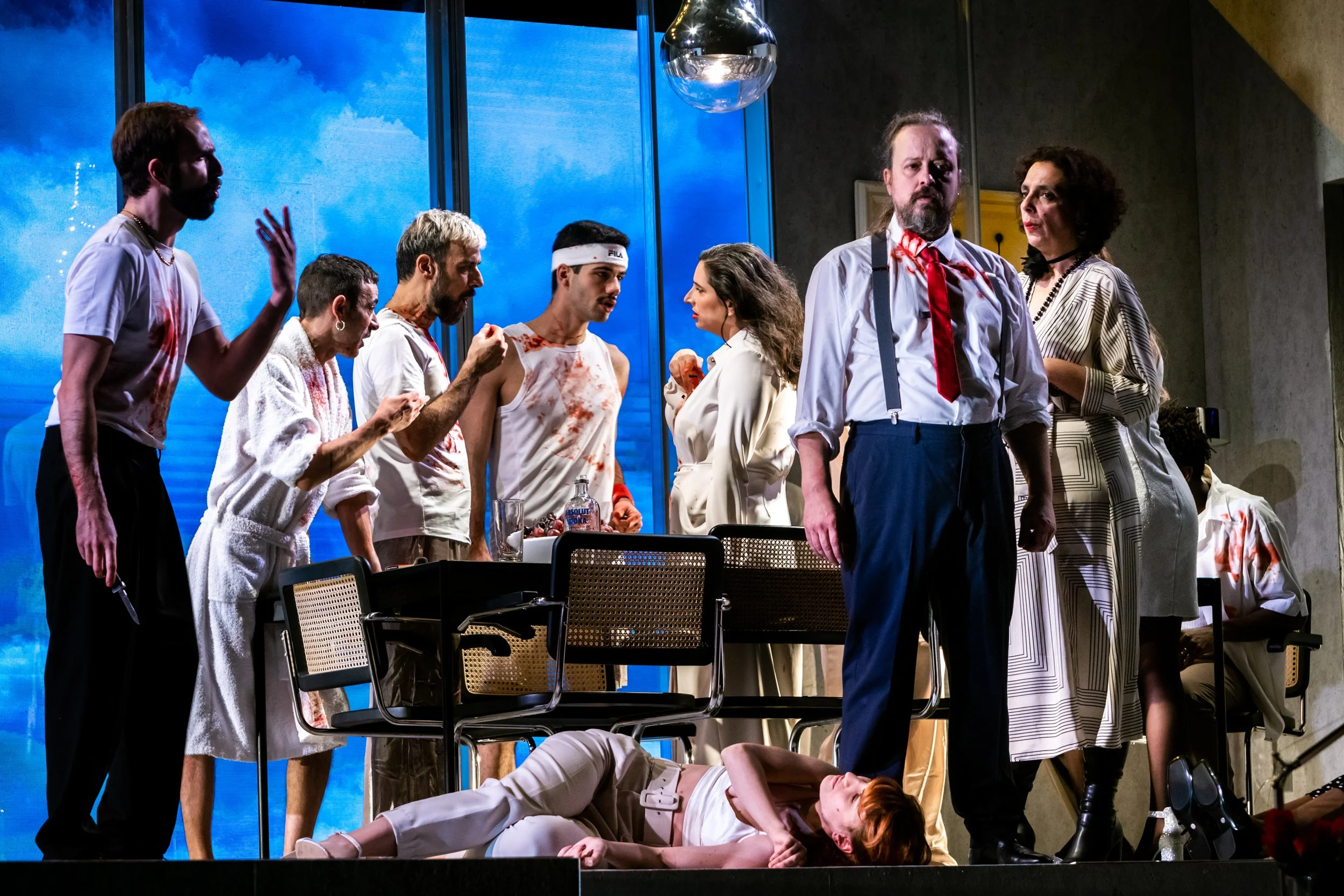 Cena de uma peça teatral com vários atores em uma cena dramática, alguns com sangue, uma pessoa deitada no chão e uma pessoa de pé com expressão séria.