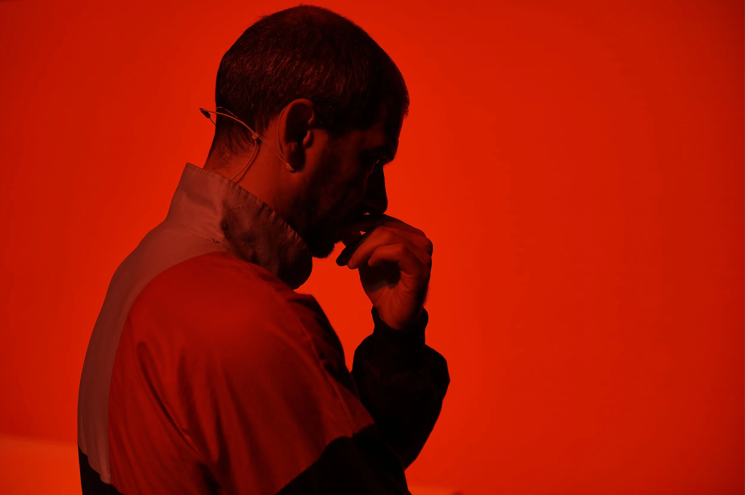 Homem pensativo com fundo vermelho, usando jaqueta e com microfone na orelha.