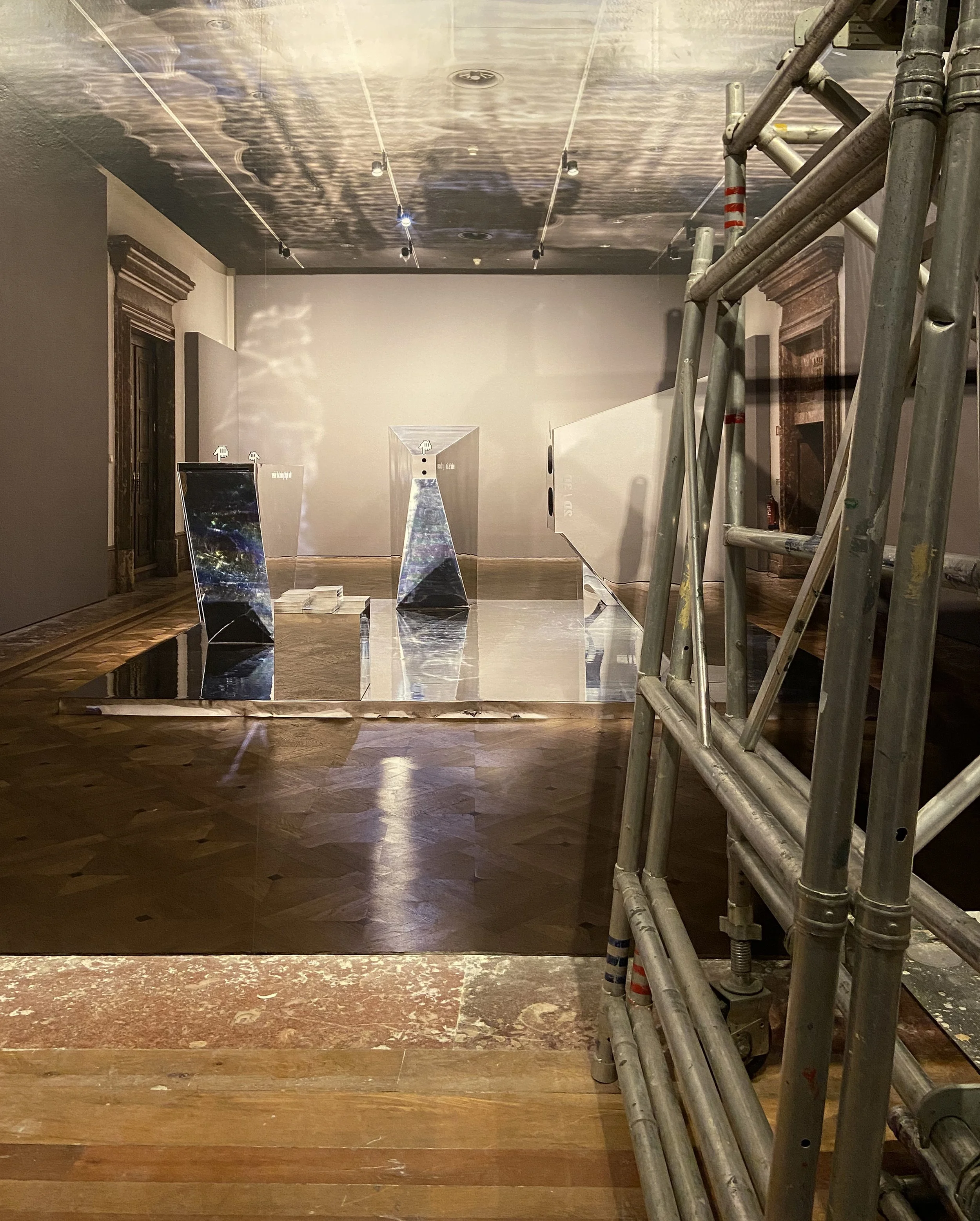 Sala de exposição ou instalação artística com estruturas de espelho e uma escada de andaime no lado direito, iluminação direcionada e piso de madeira.