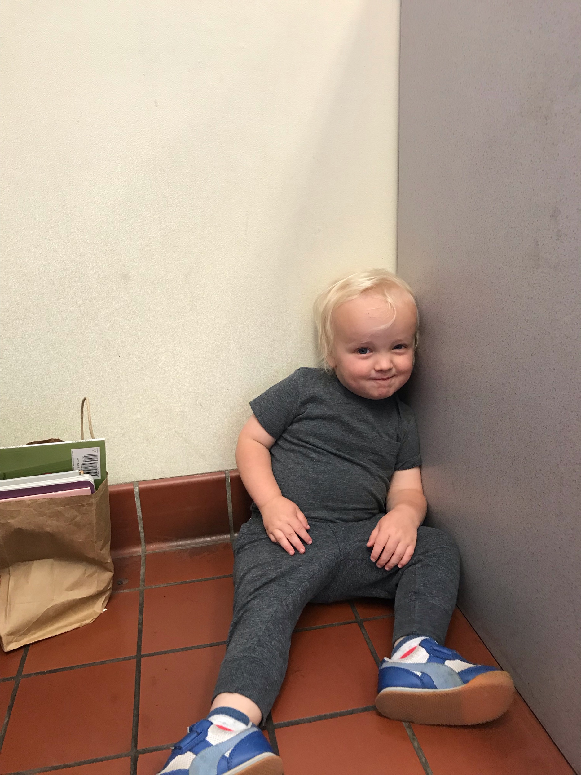 Toddler-age boy sitting in corner of post office with a bag of books. 