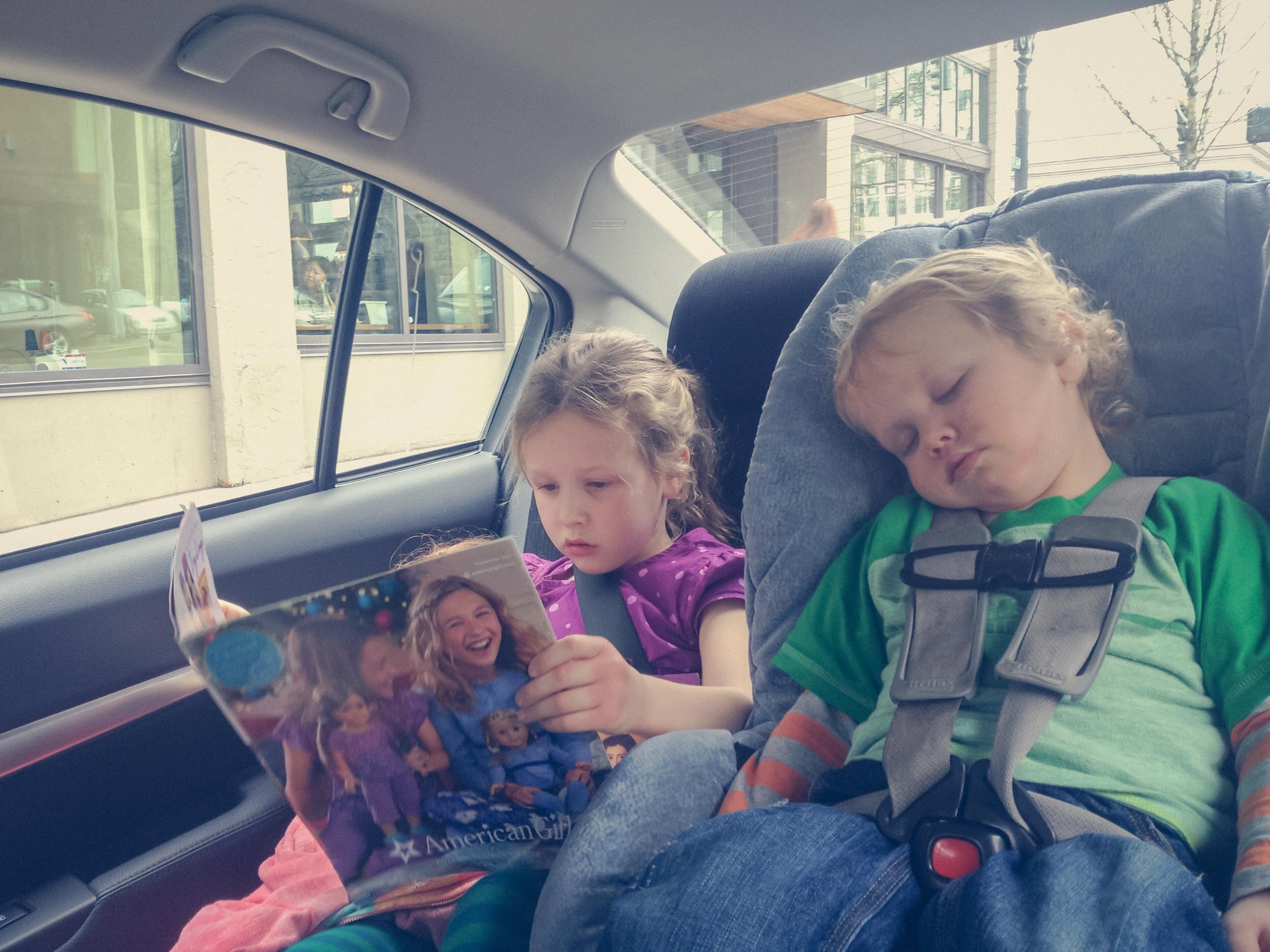 Girl reading American Girl magazine in car while little brother sleeps.
