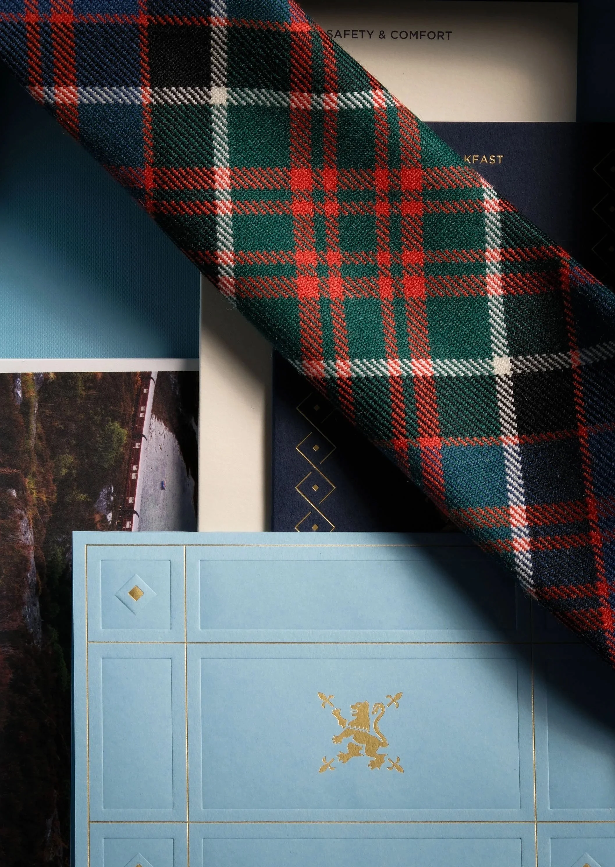 Close-up of documents, including a tartan-patterned cloth and a blue booklet with a gold lion emblem, and a printed photograph of a river with a bridge.