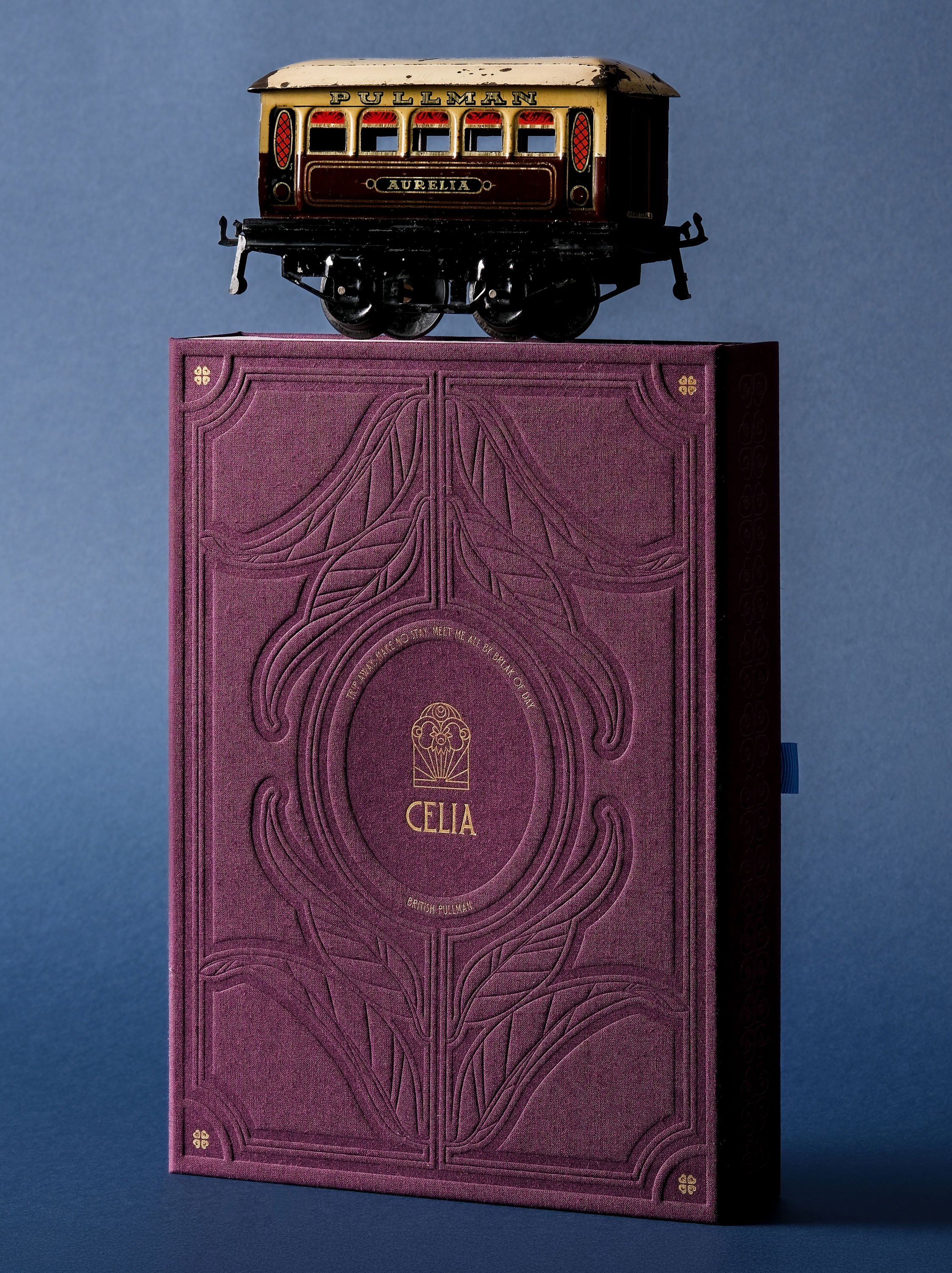A vintage tin toy train car labeled 'Pullman' and 'Aurelia' on top of a decorative pink book with embossed patterns, set against a blue background.