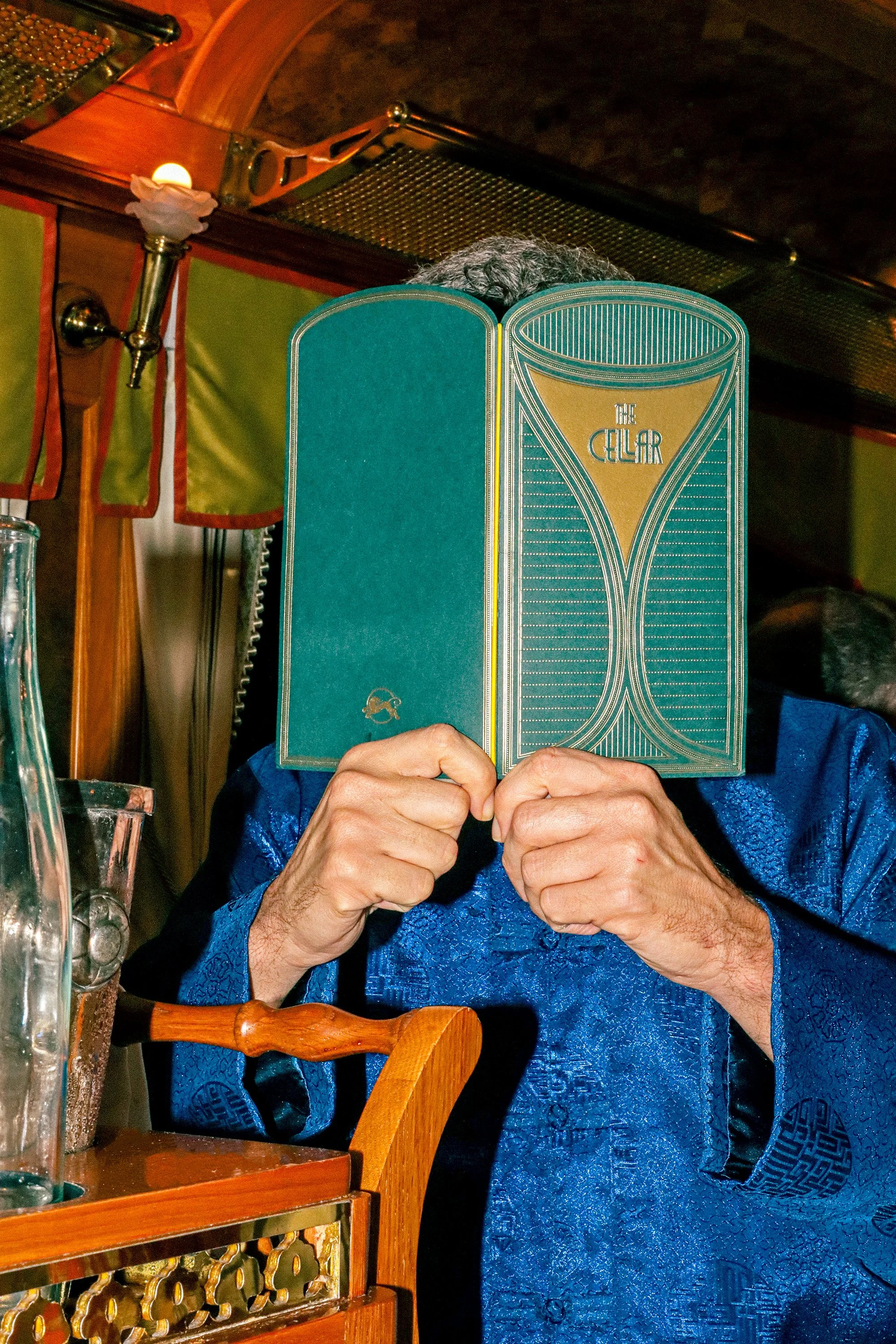 Person in a blue silk shirt holding a green menu titled “The Cellar” up to their face inside the Eastern & Oriental Express, with brass fixtures and wooden detailing in the background.