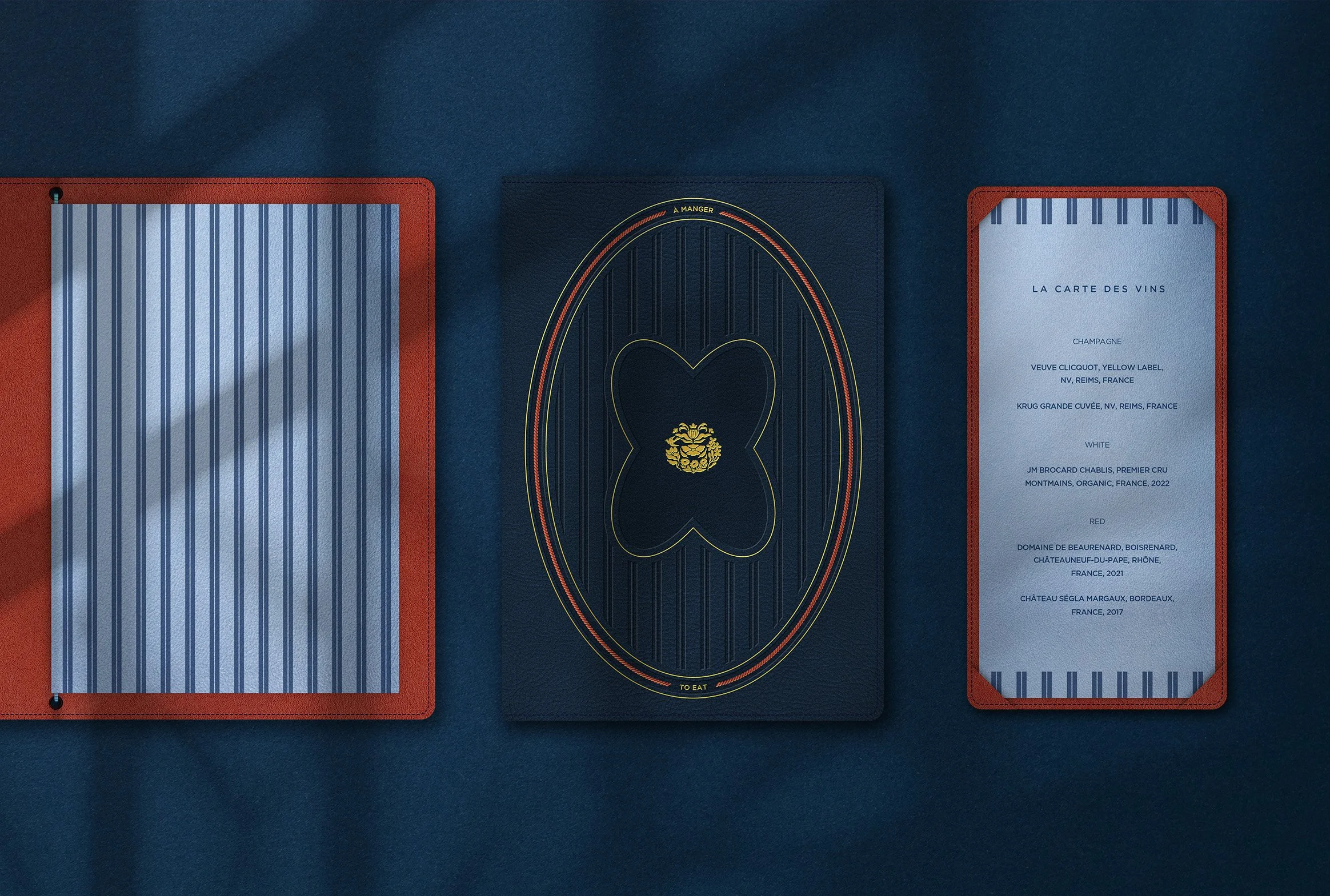 Leather oversized menu folios, one closed and one half-open, with a matching DL drinks menu alongside, set on a dark blue surface with gold and red detailing.