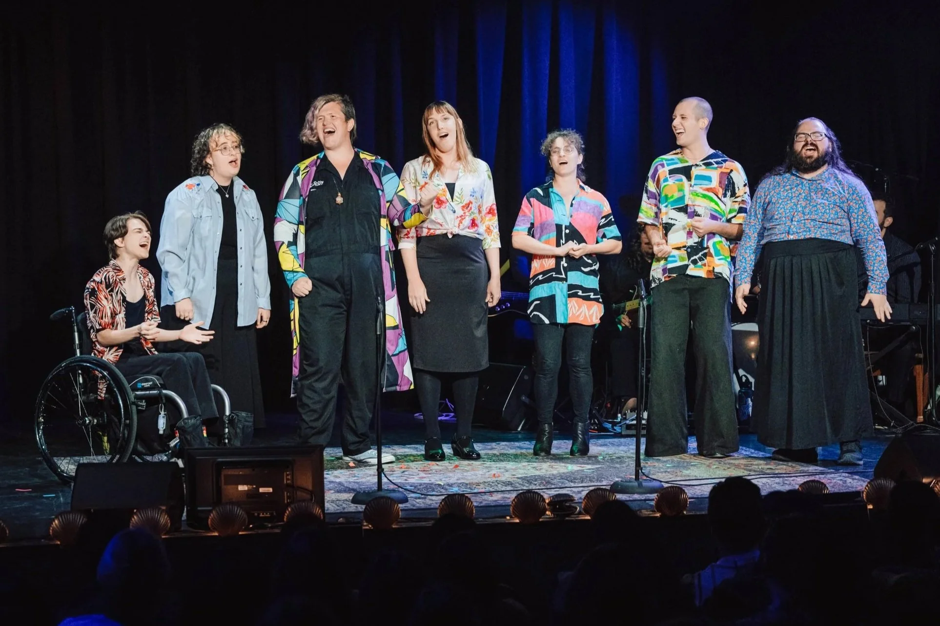 Seven trans and nonbinary vocalists dressed in colorful outfits sing with energy on stage
