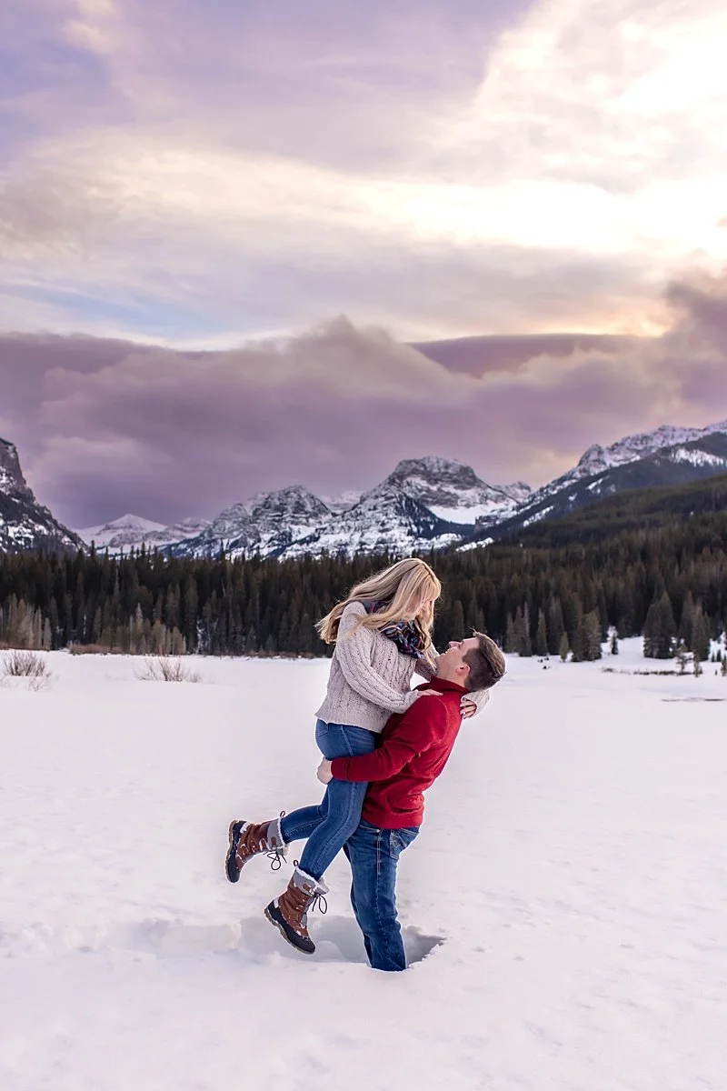 Engaged couple in the mountains, he is lifting her, they are making eye contact and smiling with sunset reflecting on the clouds behind them.